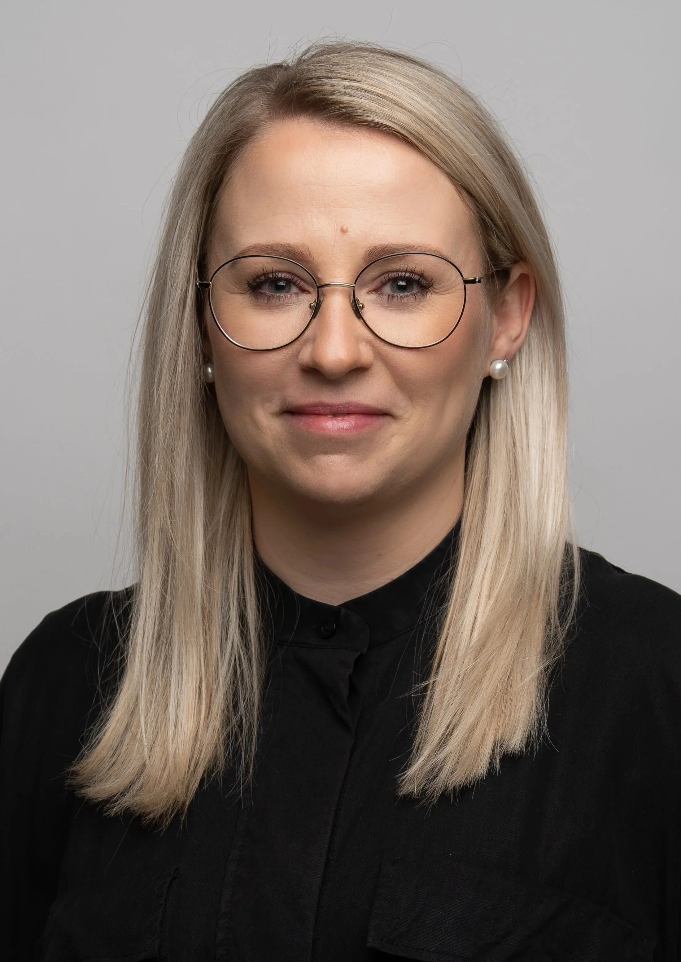 En kvinde med langt blond hår, bære store, runde brilleglas og øreringe, iført sort tøj mod en neutral grå baggrund.
