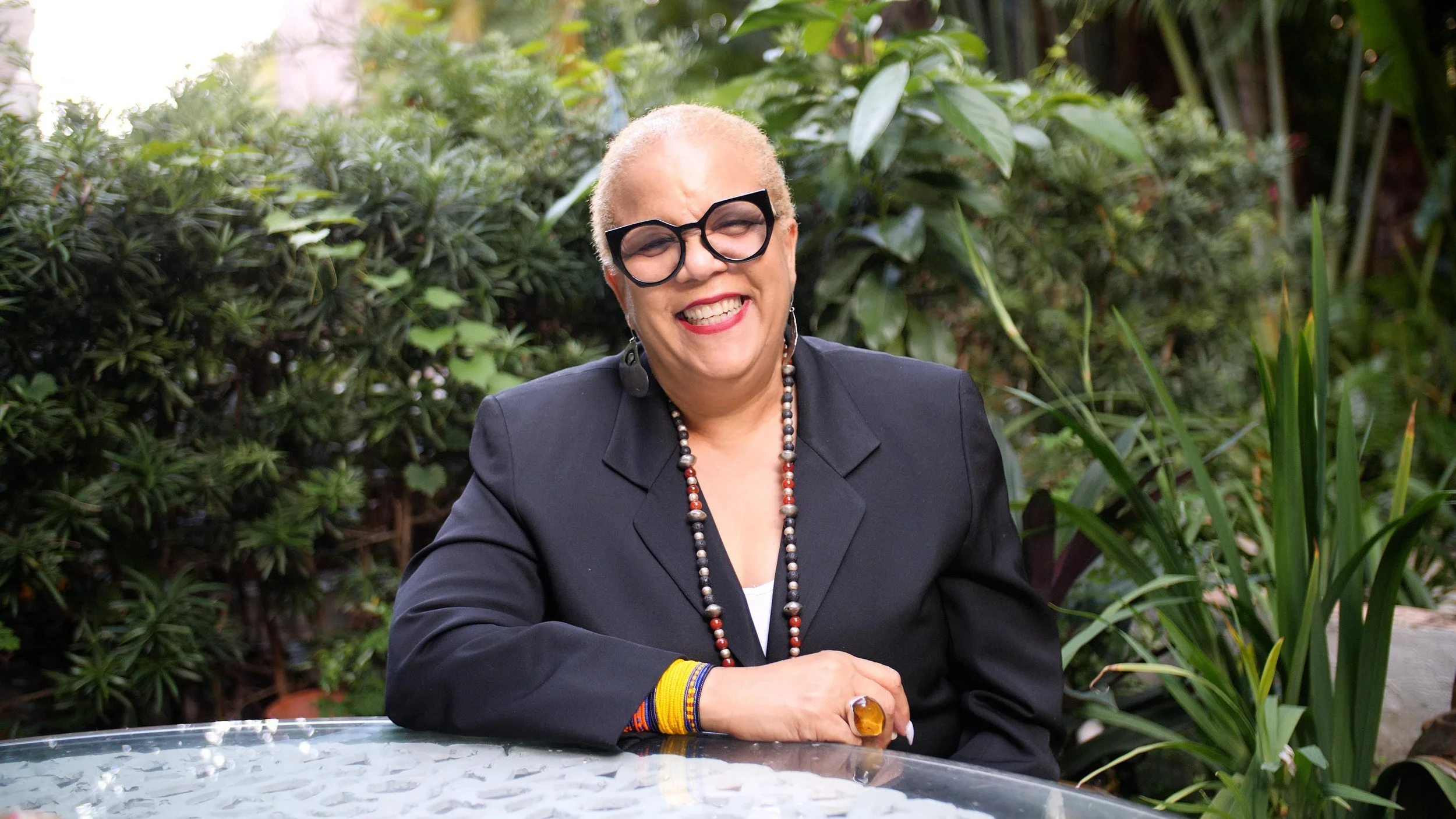 Person sitting outdoors at a glass table, wearing large black glasses and a black suit, smiling, with greenery in the background.