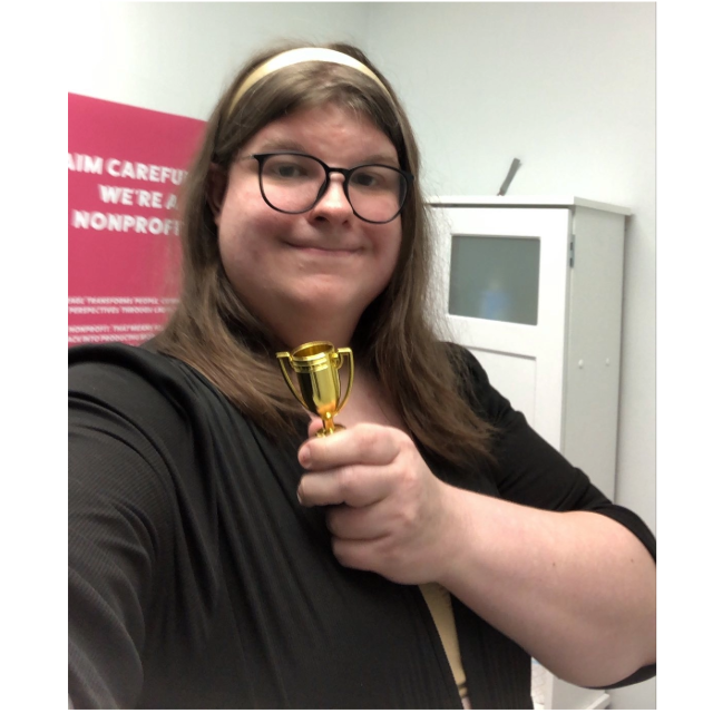 A picture of me, Billie Sainwood, a trans white woman standing in a bathroom holding a comically small trophy.