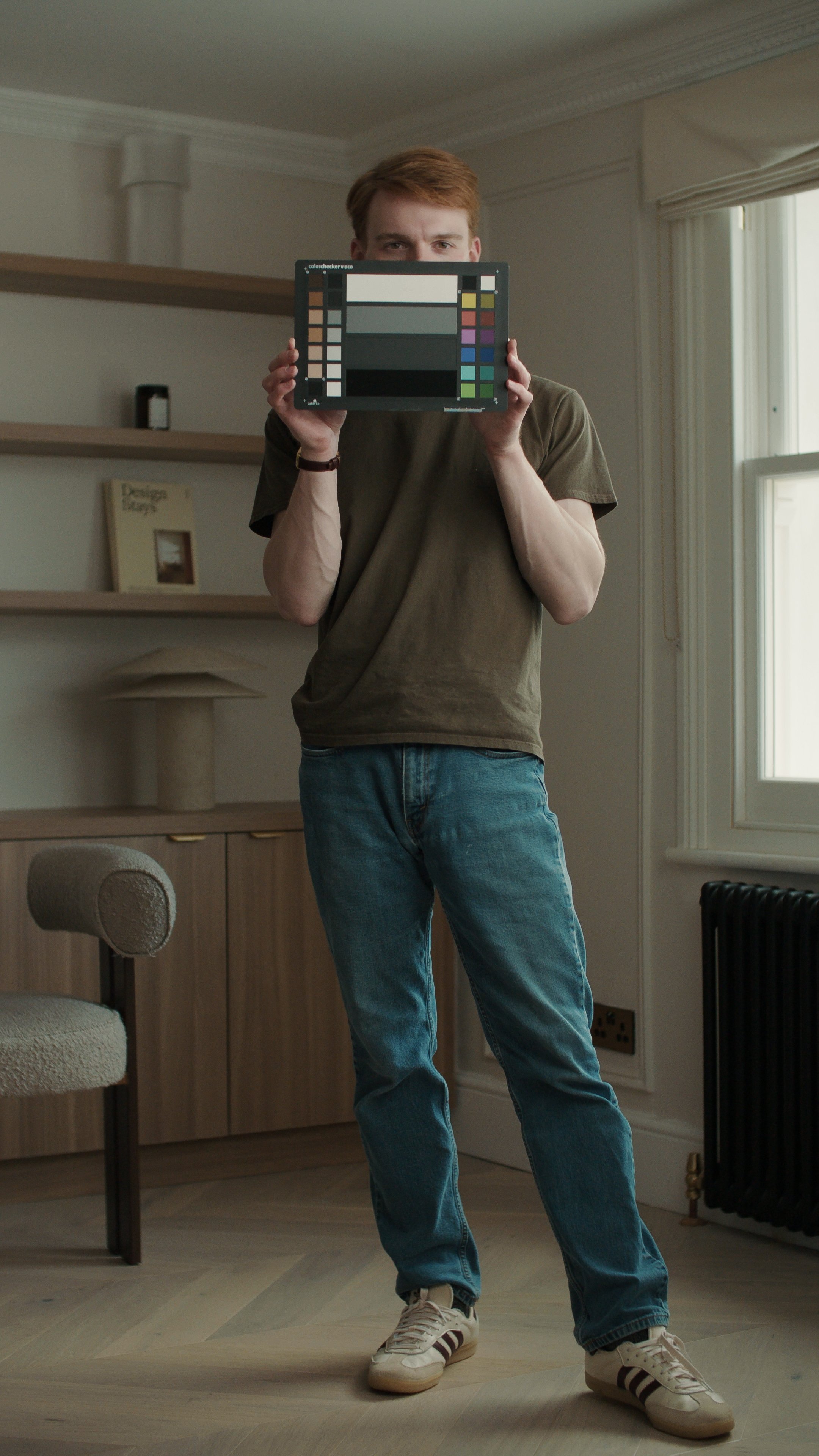 A man with reddish hair standing in a room, holding a colour calibration chart in front of his face. He is wearing a brown T-shirt, blue jeans, and sneakers, with natural light coming from a window on the right.