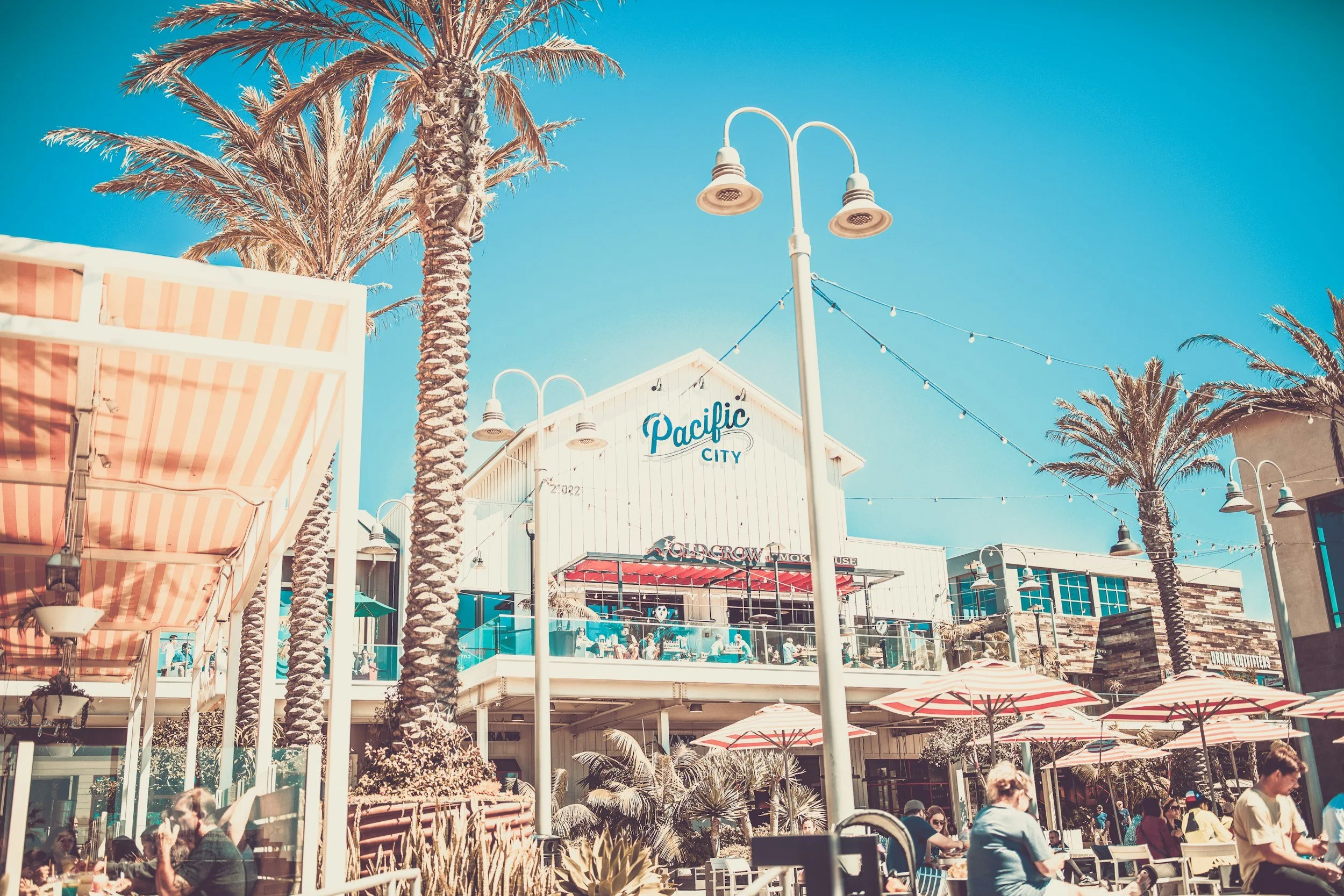 Huntington Beach outdoor shopping and dining area with tall palm trees, striped umbrellas, string lights, and a building with a sign that says 'Pacific City' under a clear blue sky.