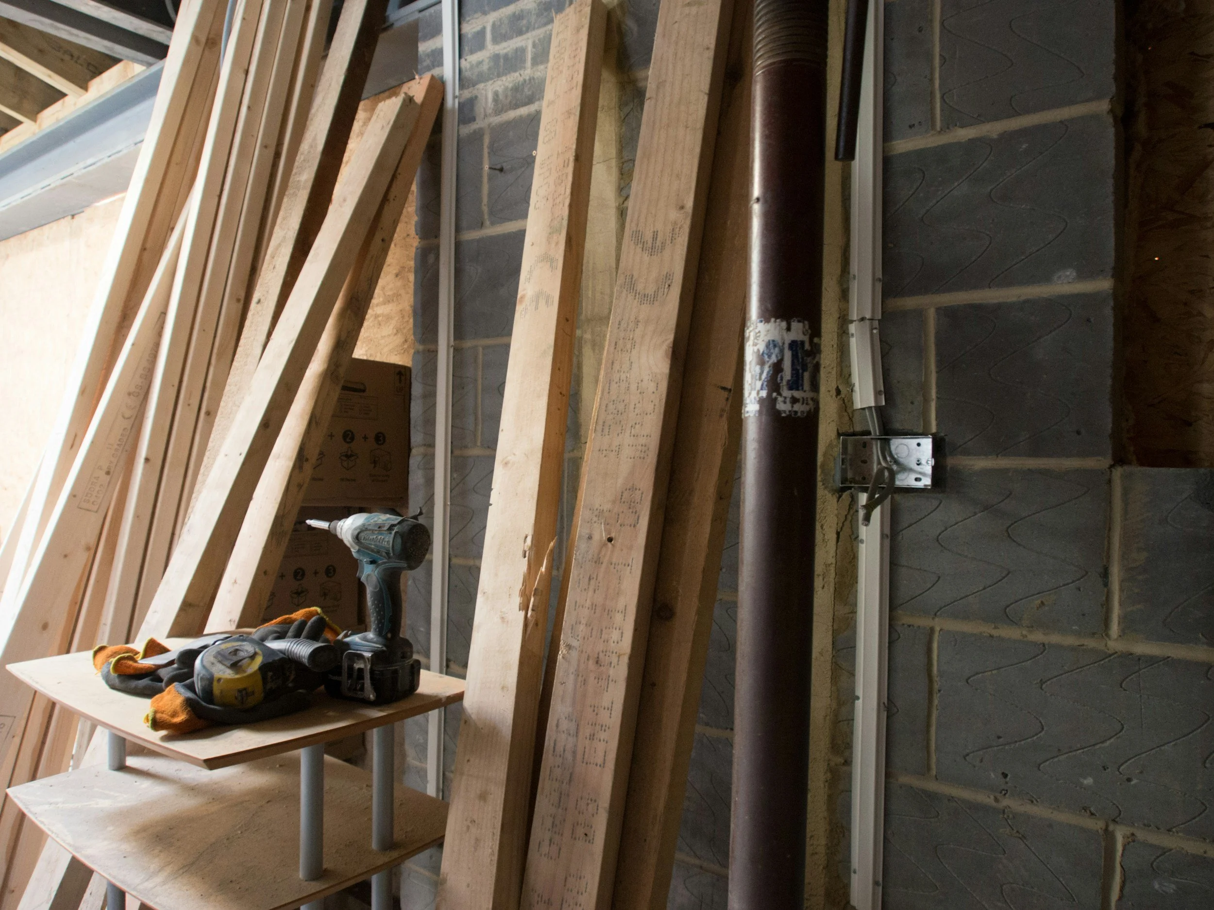 Construction tools and materials, including a cordless drill, gloves, and wood planks, are set up on a small table in front of a partially constructed wall with metal framing, a pipe, and wiring in a building under construction.
