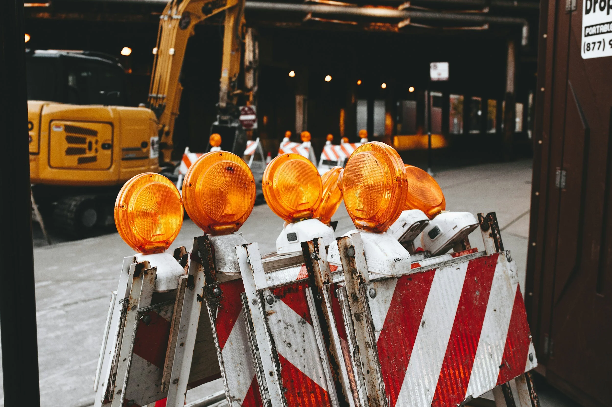 Construction site with a barricade featuring orange warning lights and a large yellow excavator in the background.