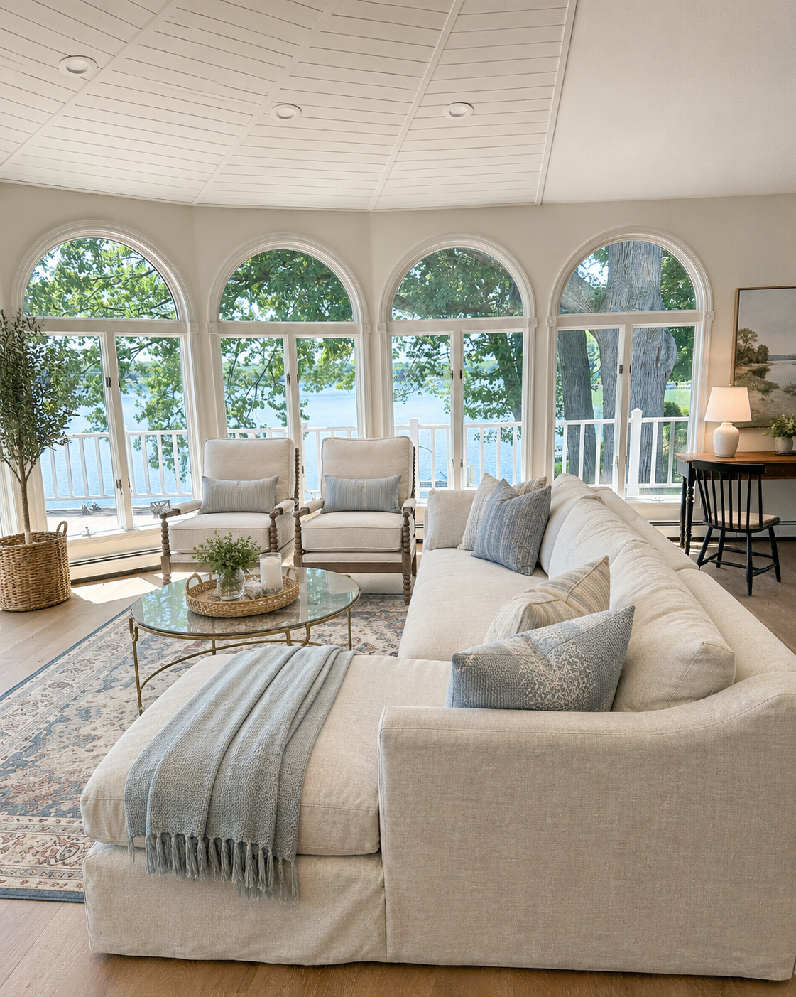 Coastal living room with large arched windows overlooking water, beige sectional sofa with blue pillows, two armchairs with pillows, round glass coffee table with plants and candles, potted plant, framed artwork, and side table with lamp.