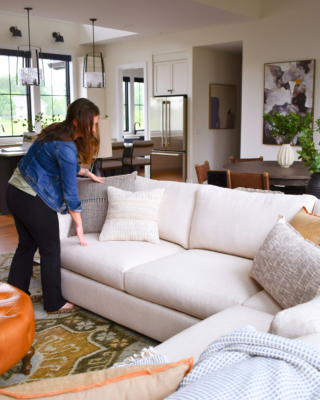 A woman arranging pillows on a white sofa in a modern living room with plants, artwork, and large windows.