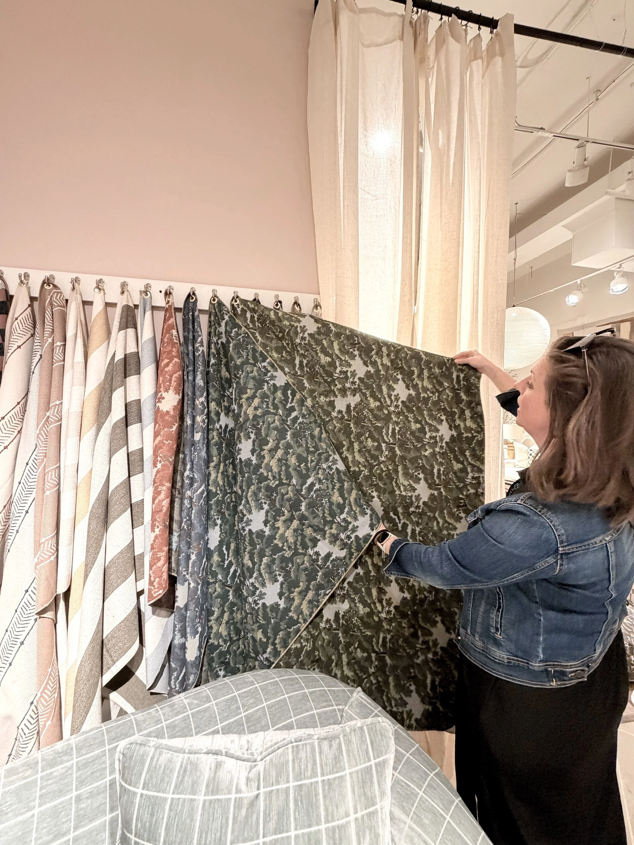 A woman wearing a denim jacket and black pants looking at upholstery fabric in an upholstery showroom, with various patterned curtains hanging on a rack in the background.