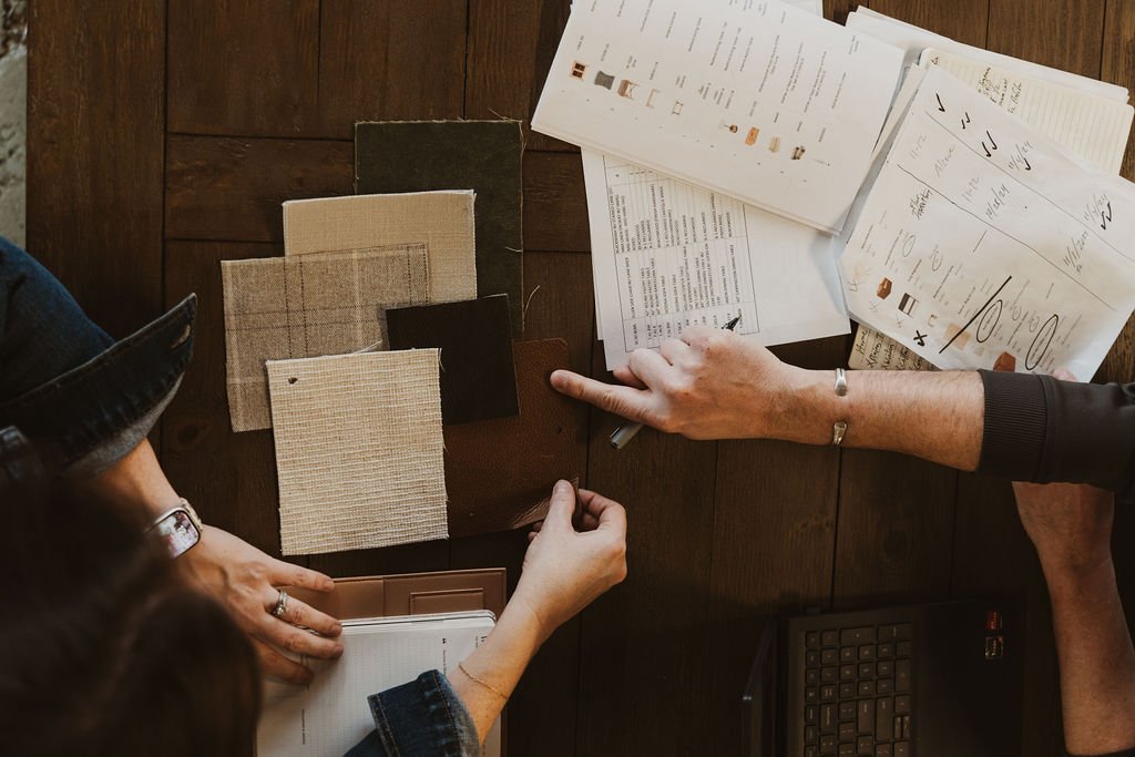 People at a wooden table reviewing fabric samples, paper swatches, and documents for a design project.