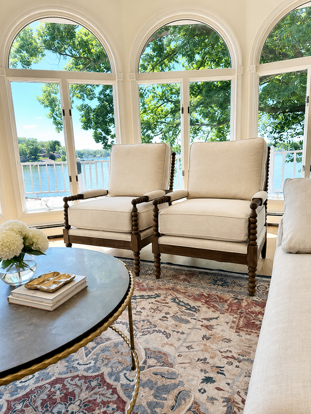 Living room with large arched windows showing a lake and green trees outside, two beige upholstered chairs with dark wooden frames, a round coffee table with a floral arrangement and books, and a patterned rug.