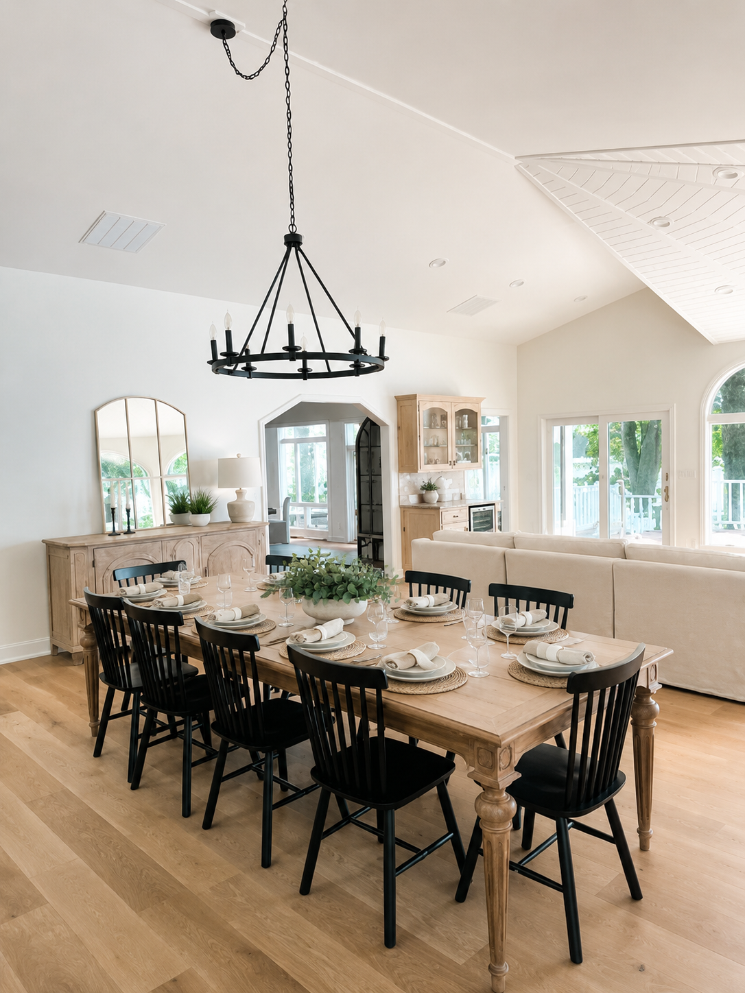 A bright dining room with a large wooden table set for a meal, surrounded by black chairs, and a farmhouse chandelier hanging overhead. There are windows and a sliding glass door that let in natural light, and a sideboard with plants and a mirror.