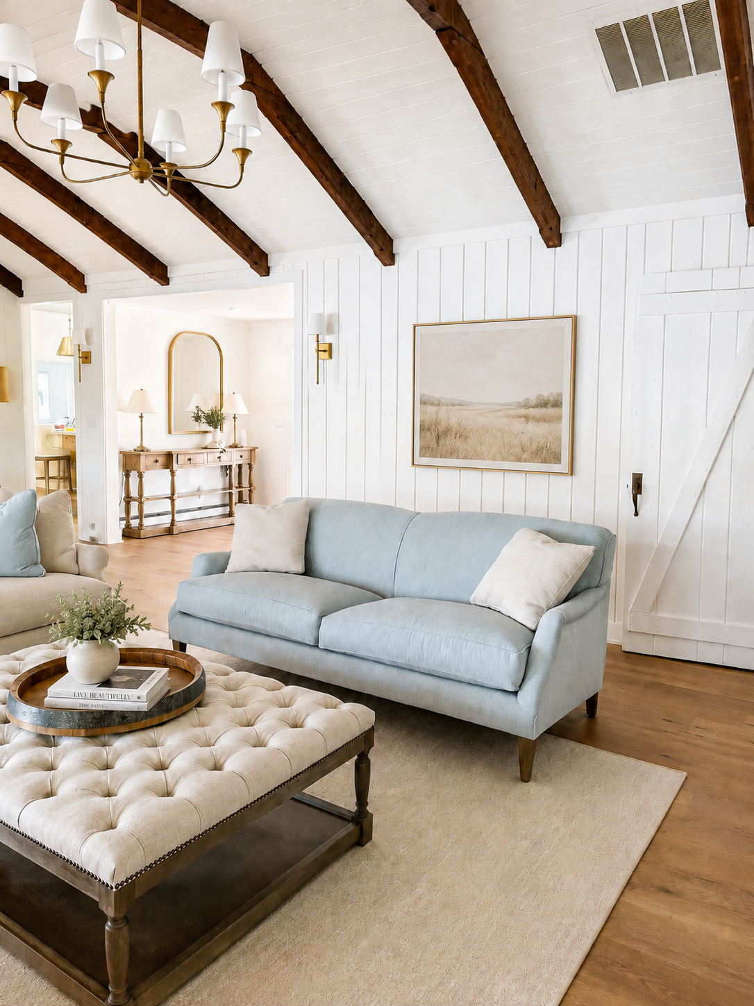 A living room with a vaulted white ceiling with dark wood beams, a large blue sofa with pillows, a tufted beige ottoman with a tray and a vase, and a wall art of a field landscape. There's a doorway leading to a hallway with a console table, mirror, 