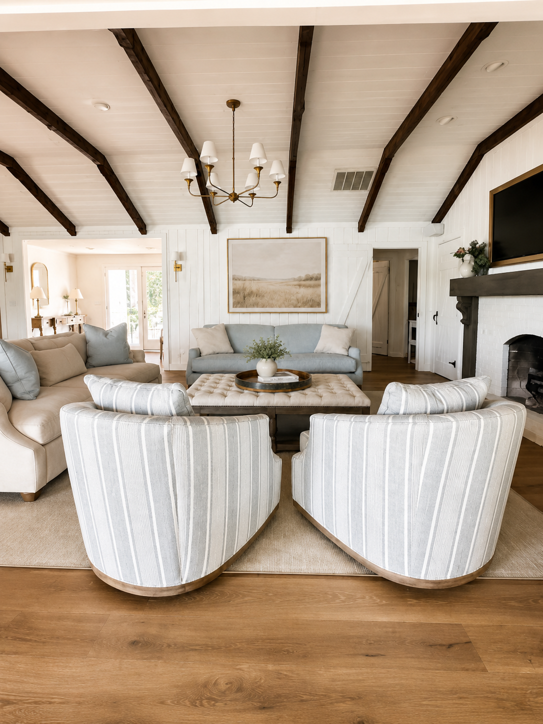 Living room with white shiplap walls and dark wooden ceiling beams, featuring a chandelier, a fireplace with a dark mantel and TV, and a seating area with various beige and light blue sofas and striped armchairs around a round coffee table with a pla
