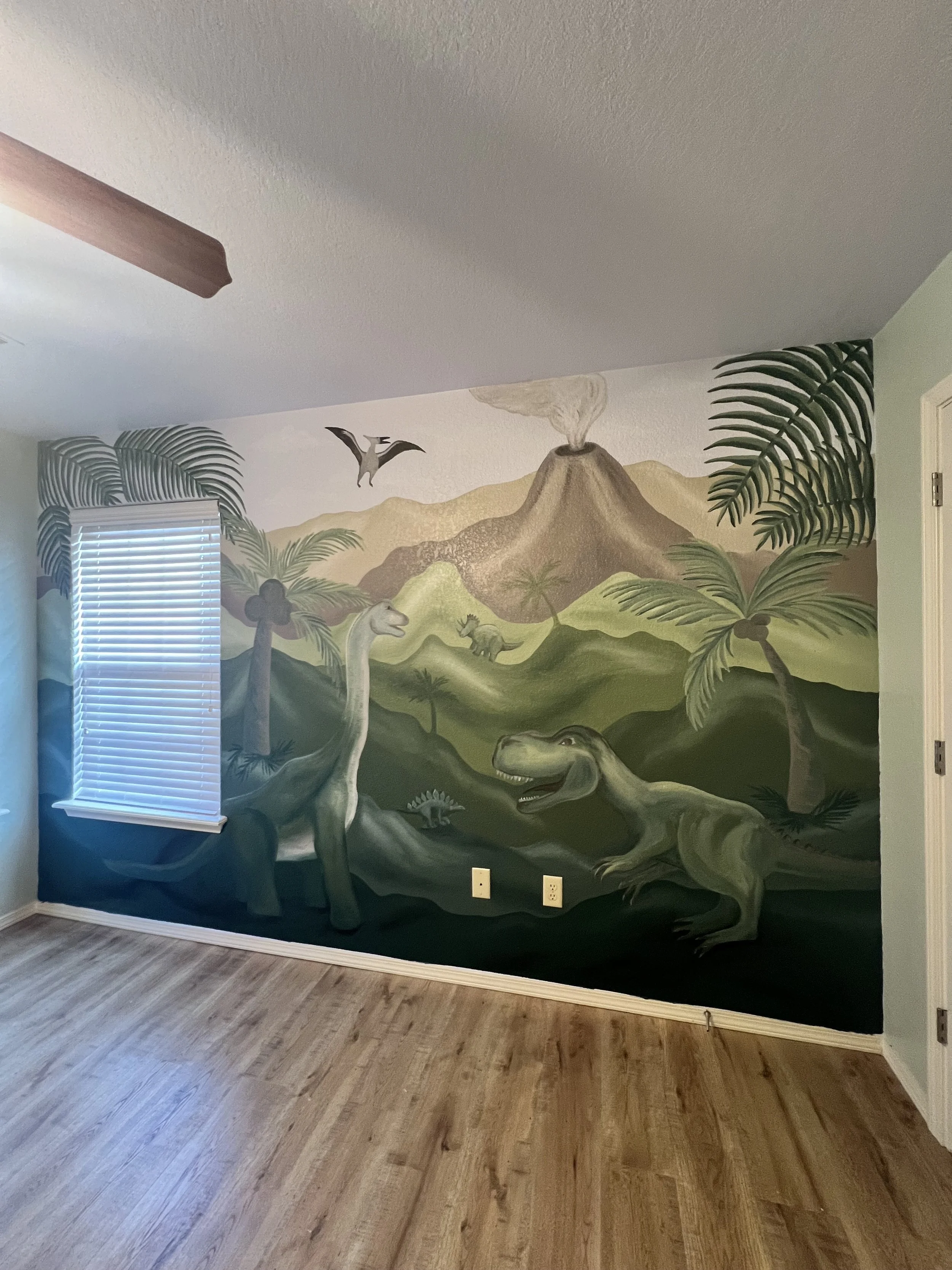 A wall mural depicting a prehistoric scene with dinosaurs, lush green plants, palm trees, a volcano erupting, and flying pterodactyls, in a bright room with wooden flooring.