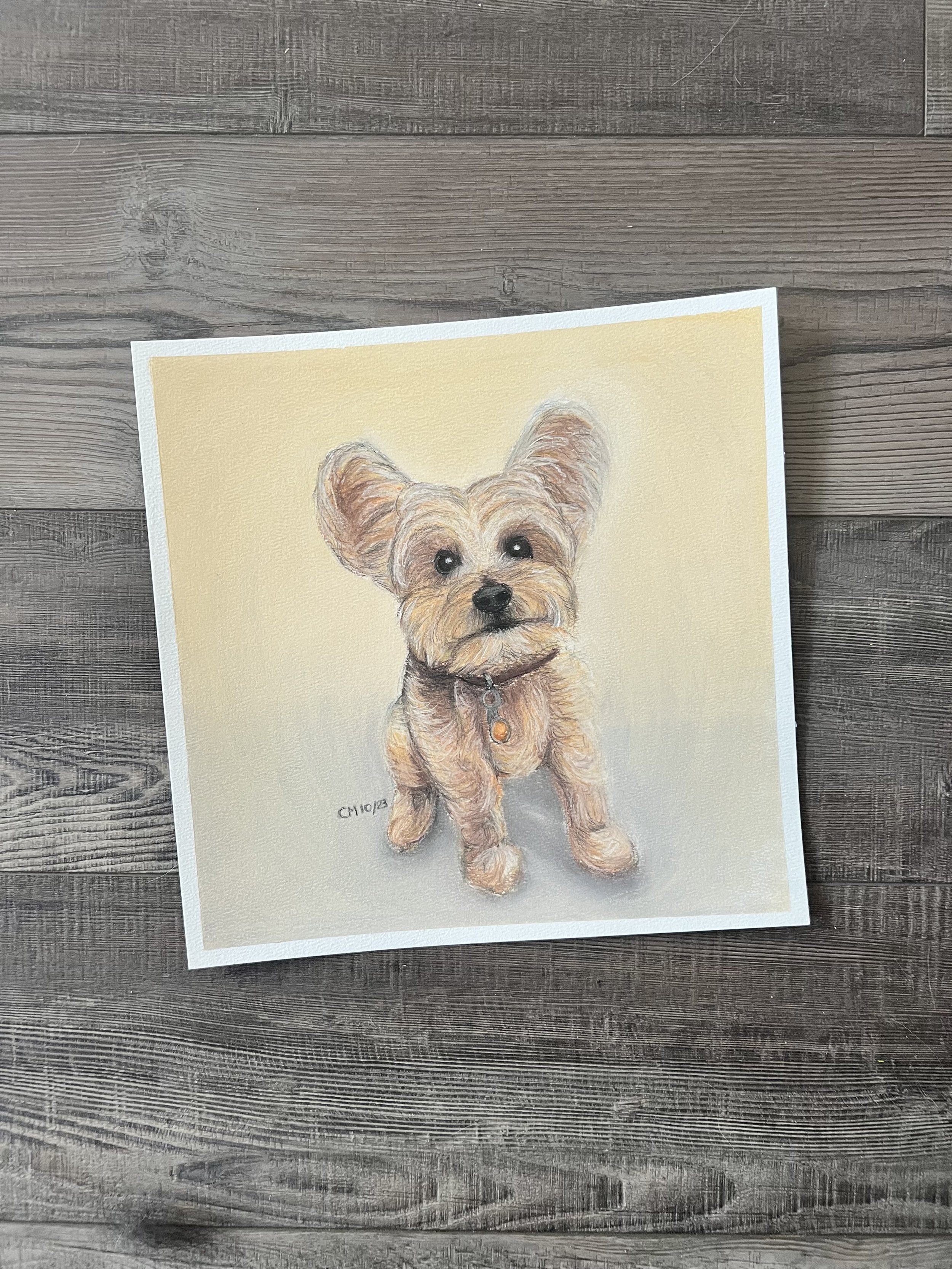 Painting of a small tan dog with large ears, black eyes, and a black nose, wearing a collar with a tag, on a light beige background, on a wooden surface.