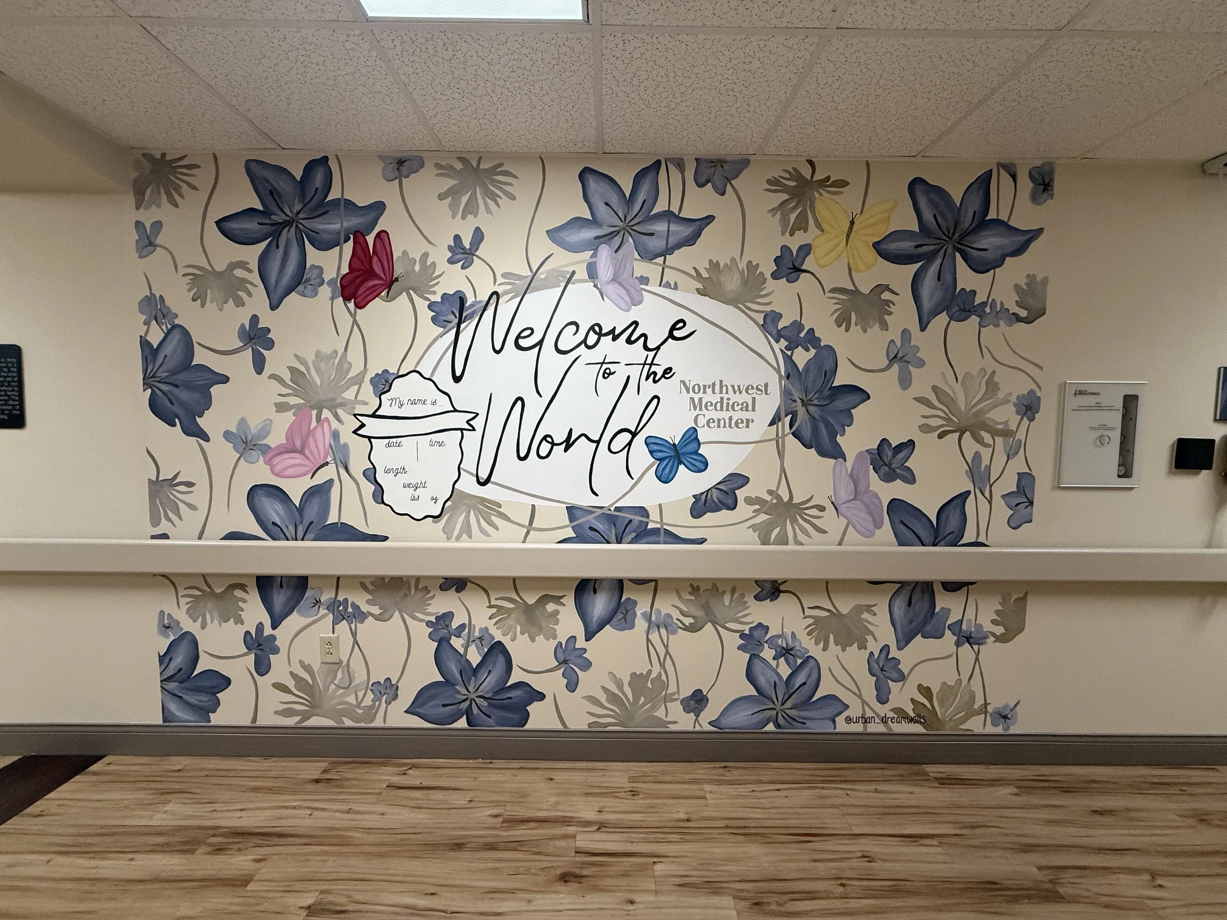 Decorative wall with floral mural and Welcome sign for Northwest Medical Center, featuring blue, purple, yellow, and pink flowers, with a small white board on the left and a white electrical outlet and panel on the right.