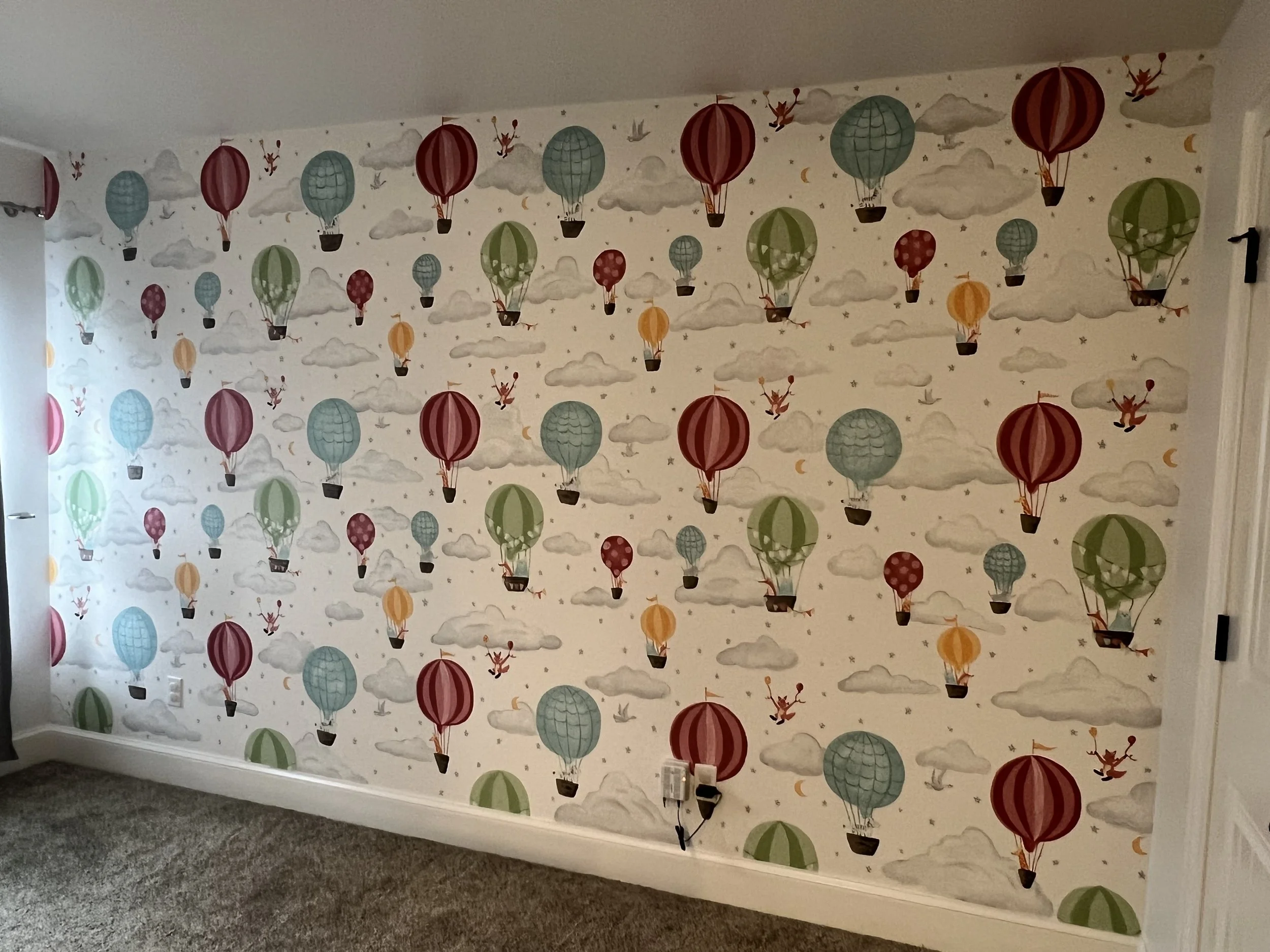 Wall with colorful hot air balloon and cloud wallpaper pattern in a child's room, with electrical outlets and a gray carpeted floor.