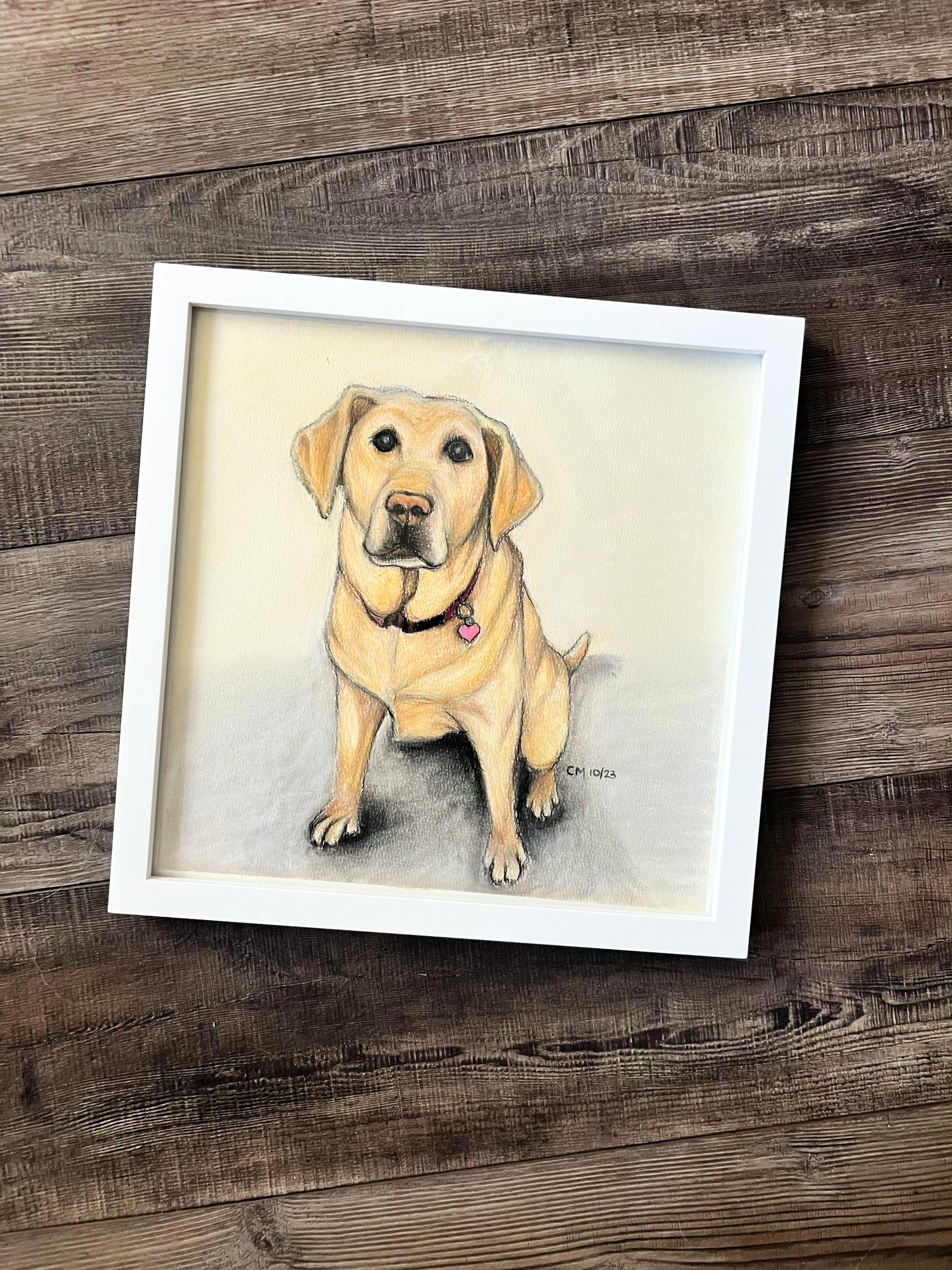 A framed watercolor painting of a yellow Labrador retriever dog with black eyes, a black nose, and a pink heart-shaped tag on its collar, sitting on a textured surface against a light background.