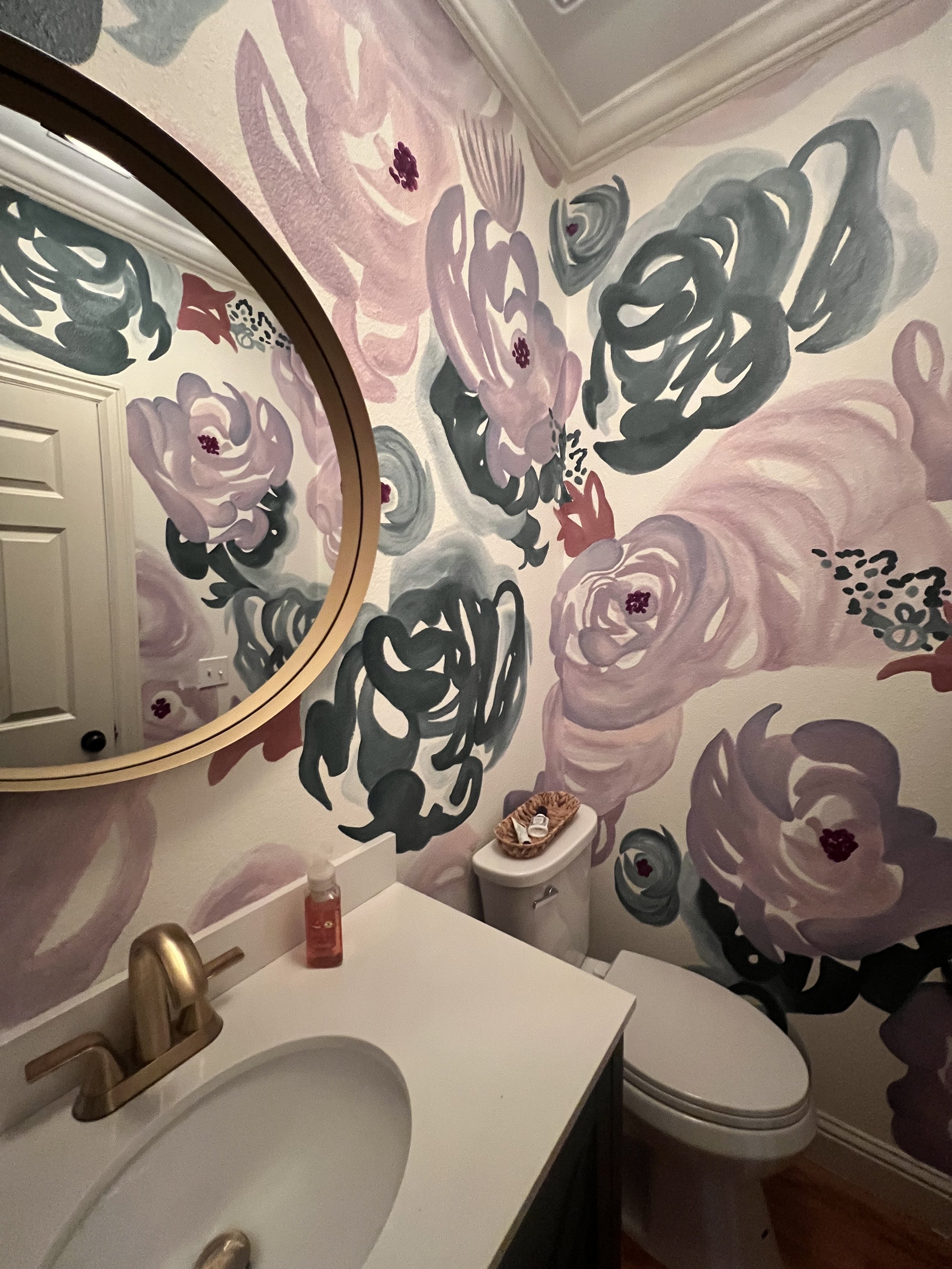 Bathroom with a floral mural wall in shades of pink, purple, black, and gray. A round mirror with a gold frame, a white countertop with a gold faucet, a soap dispenser, and a toilet with a basket on top.