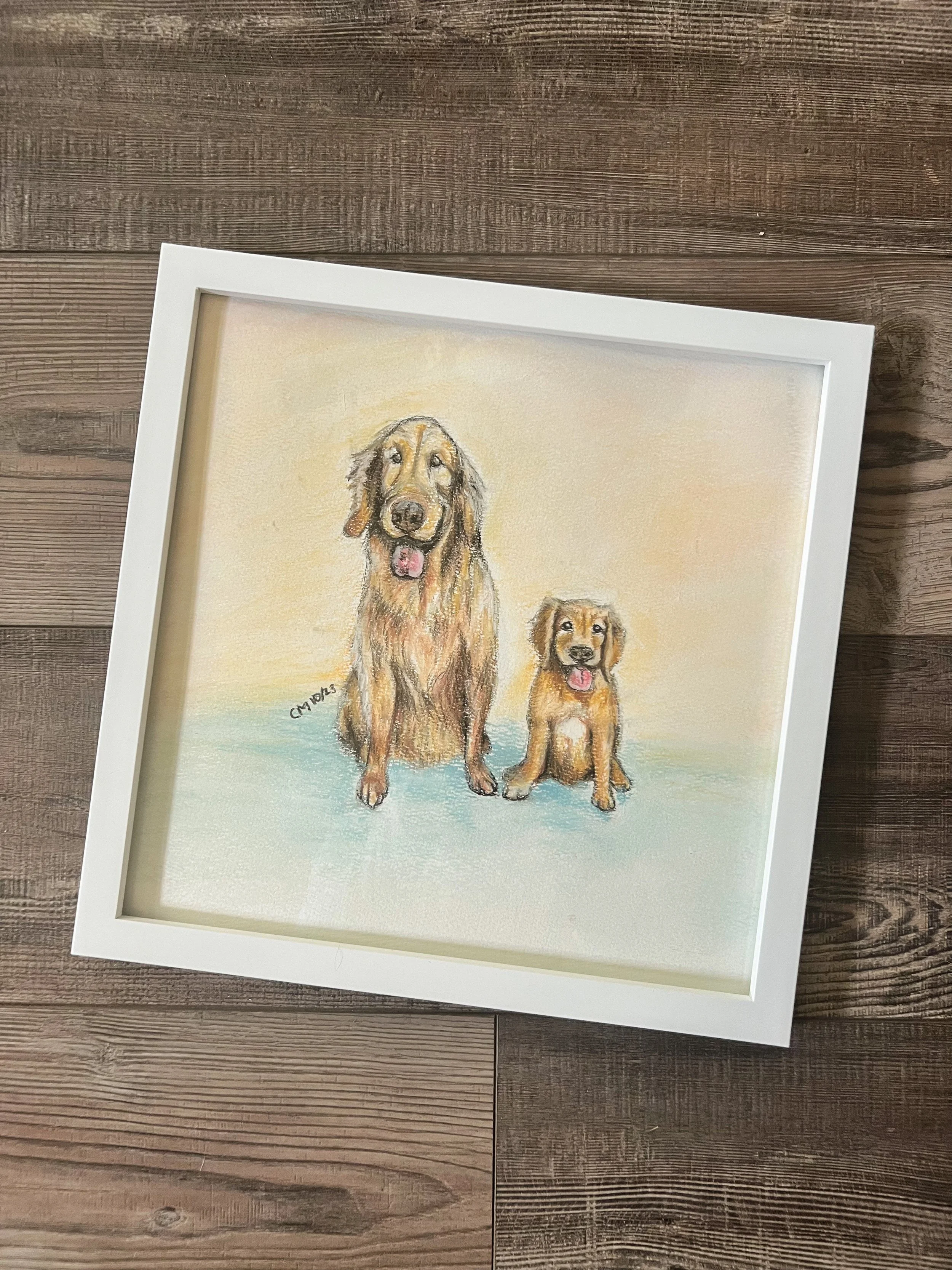 A framed pastel or watercolor painting of two golden retriever dogs sitting on a light-colored background, on a wooden floor.