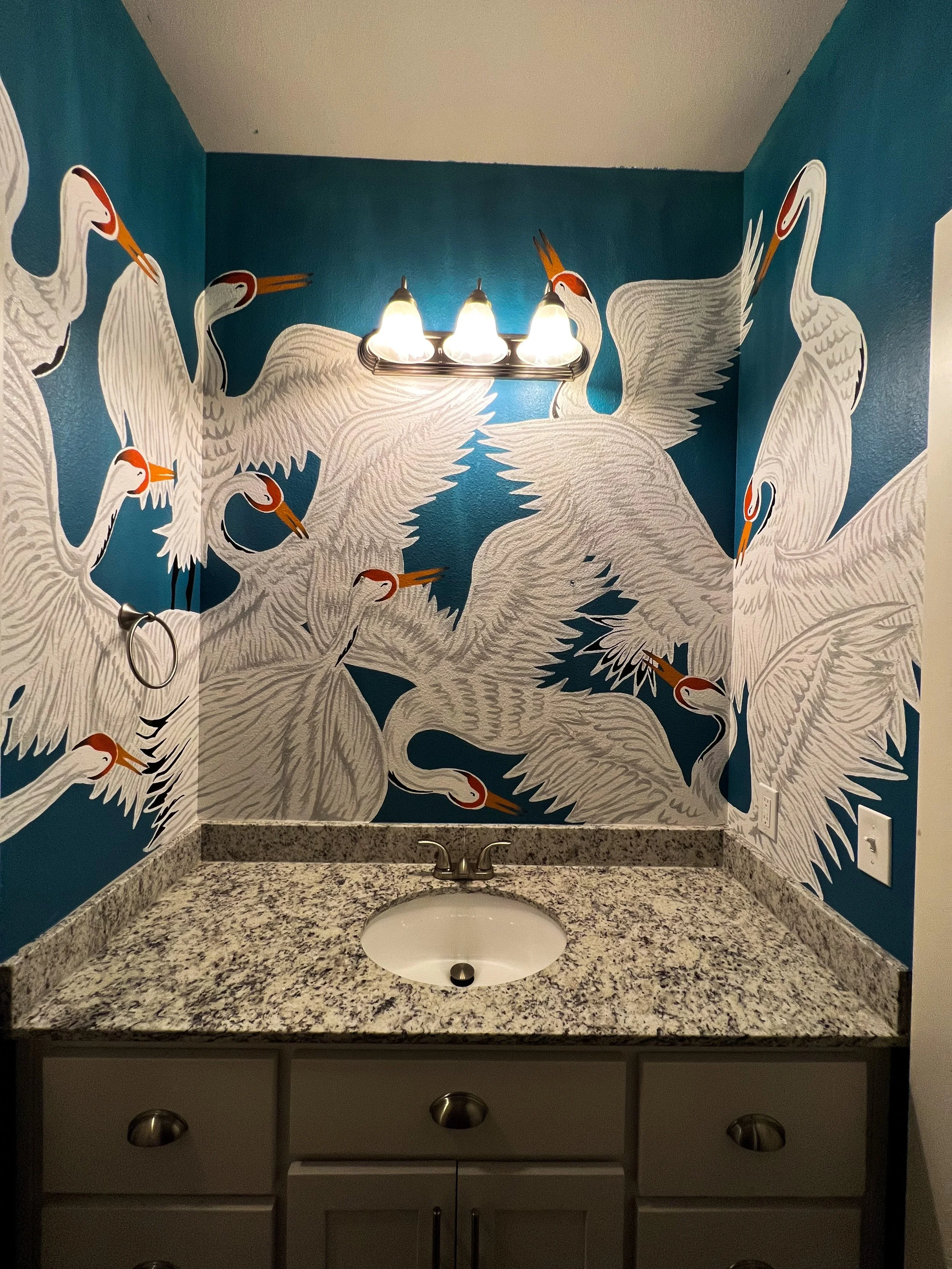 Bathroom vanity with a granite countertop, under-mount sink, and a wall mural of white cranes on a blue background, illuminated by a three-bulb light fixture.