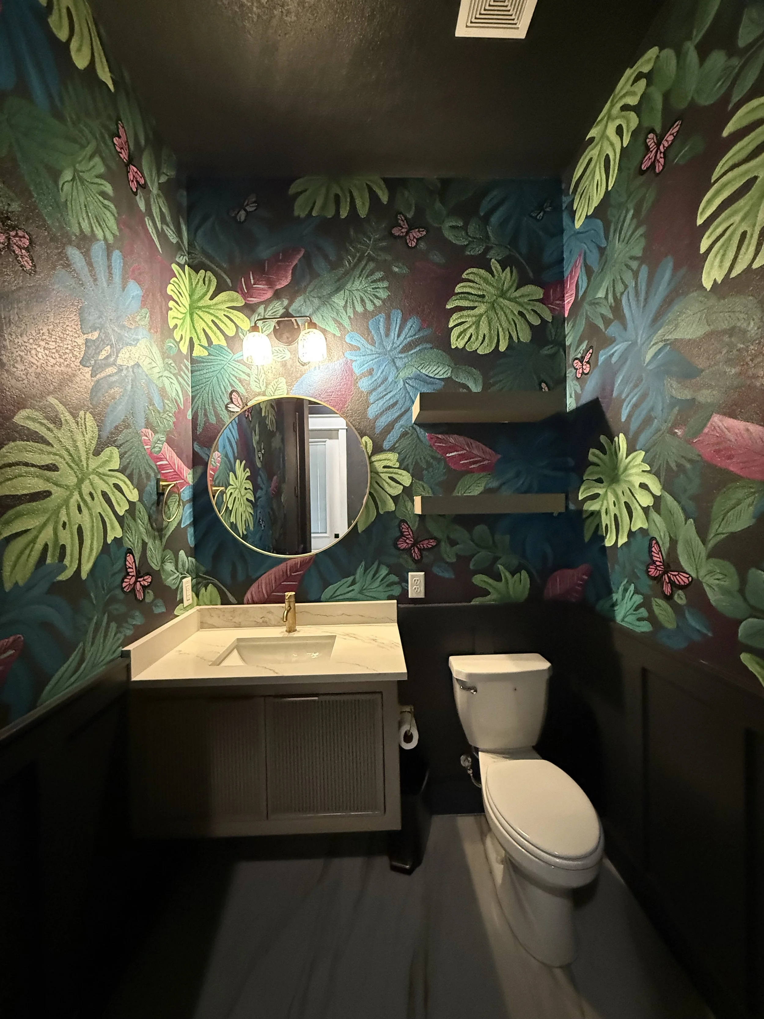 Small bathroom with dark tropical leaf and butterfly patterned wallpaper, round mirror, marble countertop sink with gold faucet, toilet, and wall shelves.