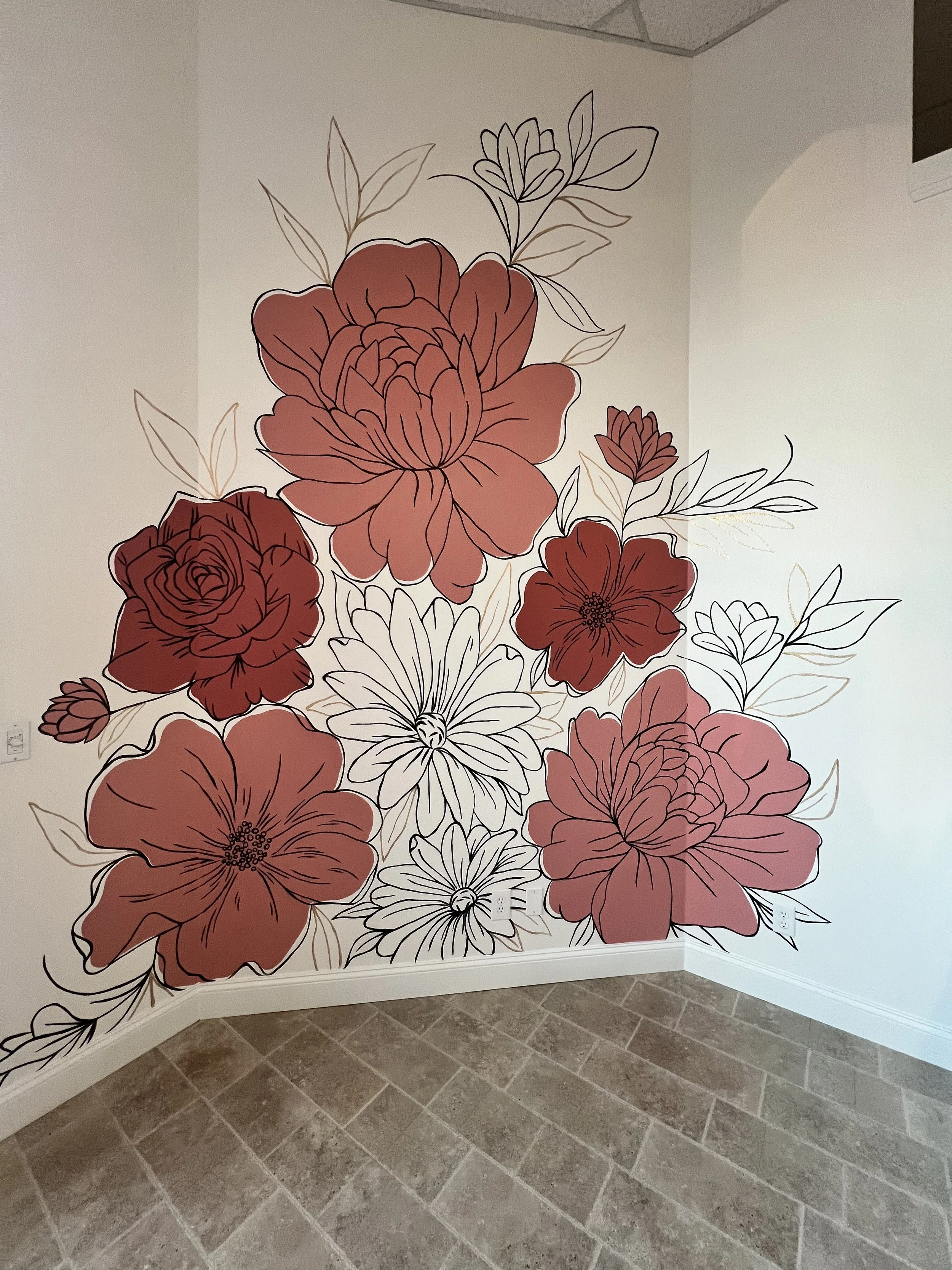 Wall mural of large pink and red flowers with leaves, outlined in black with some gold accents, on a white wall in a room with tiled floor.