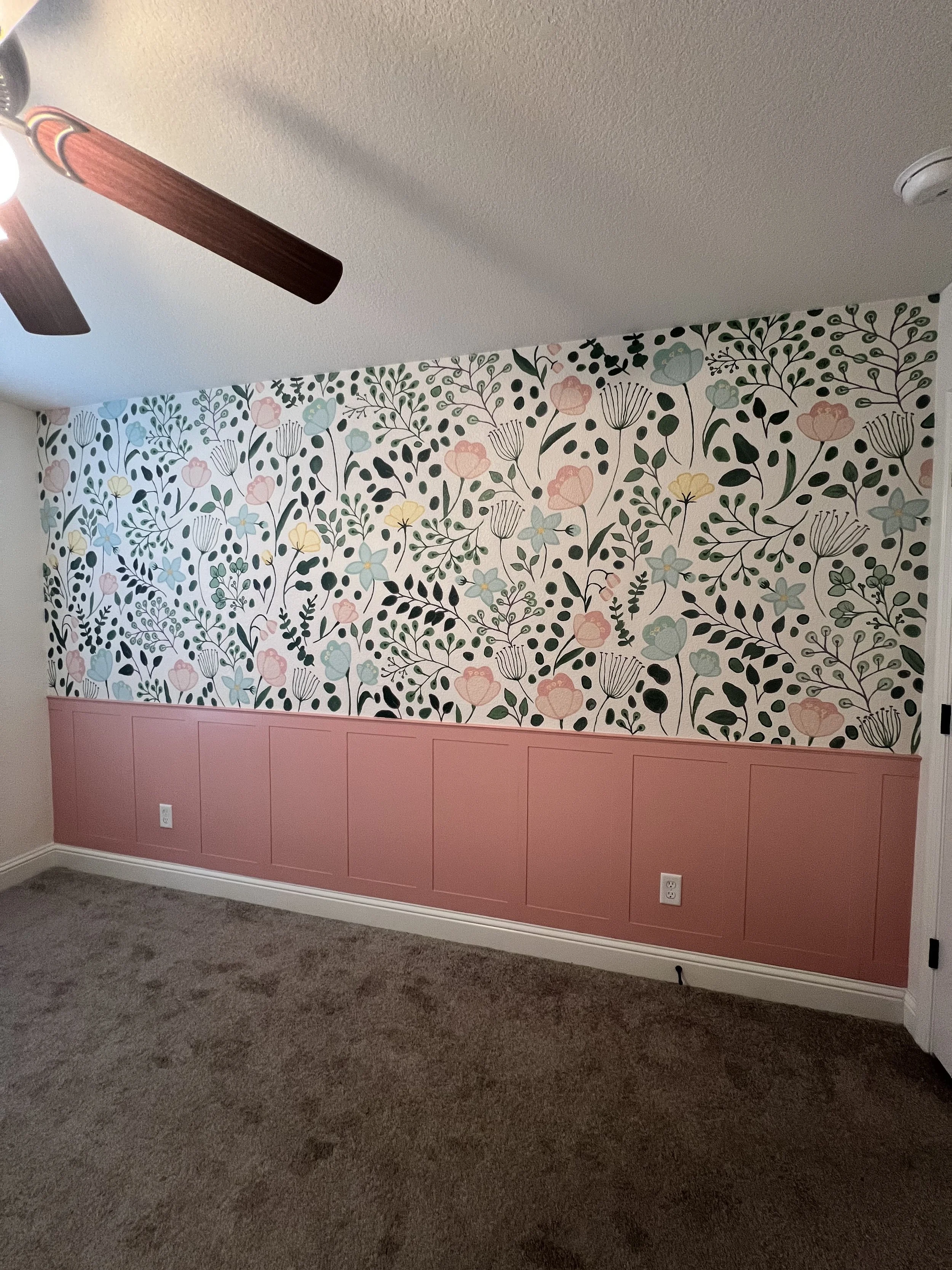 A room with a floral wallpapered wall featuring pink, blue, yellow, and green flowers and leaves, topped with a pink wainscoting. The room has a ceiling fan with dark blades and a light, and beige carpet.