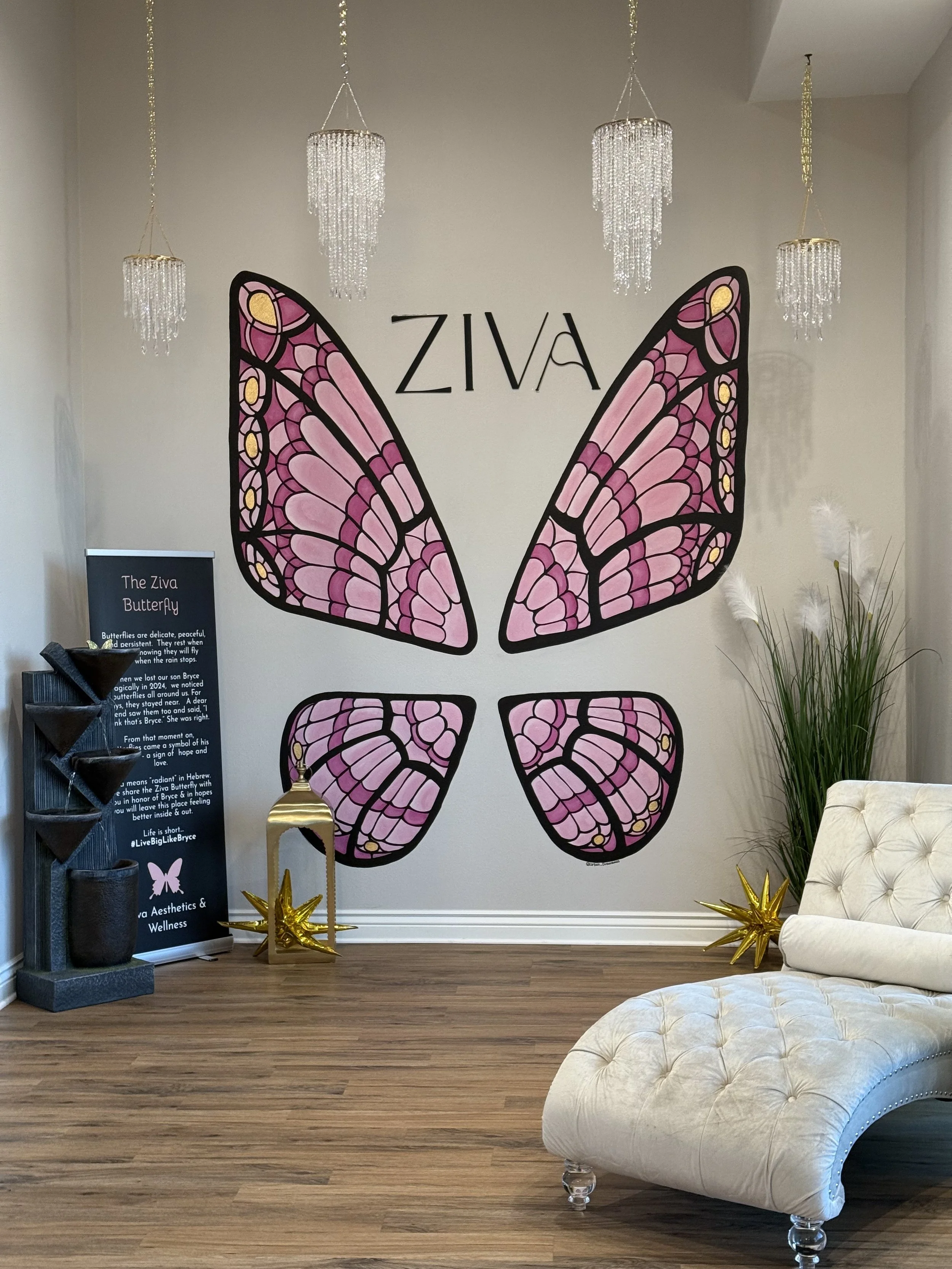 Decorative interior space with a large butterfly wall art made of pink stained glass, the word 'ZIVA' above it, chandelier lights hanging from the ceiling, a white tufted chaise lounge, a tall green plant, and a black and gold display sign.