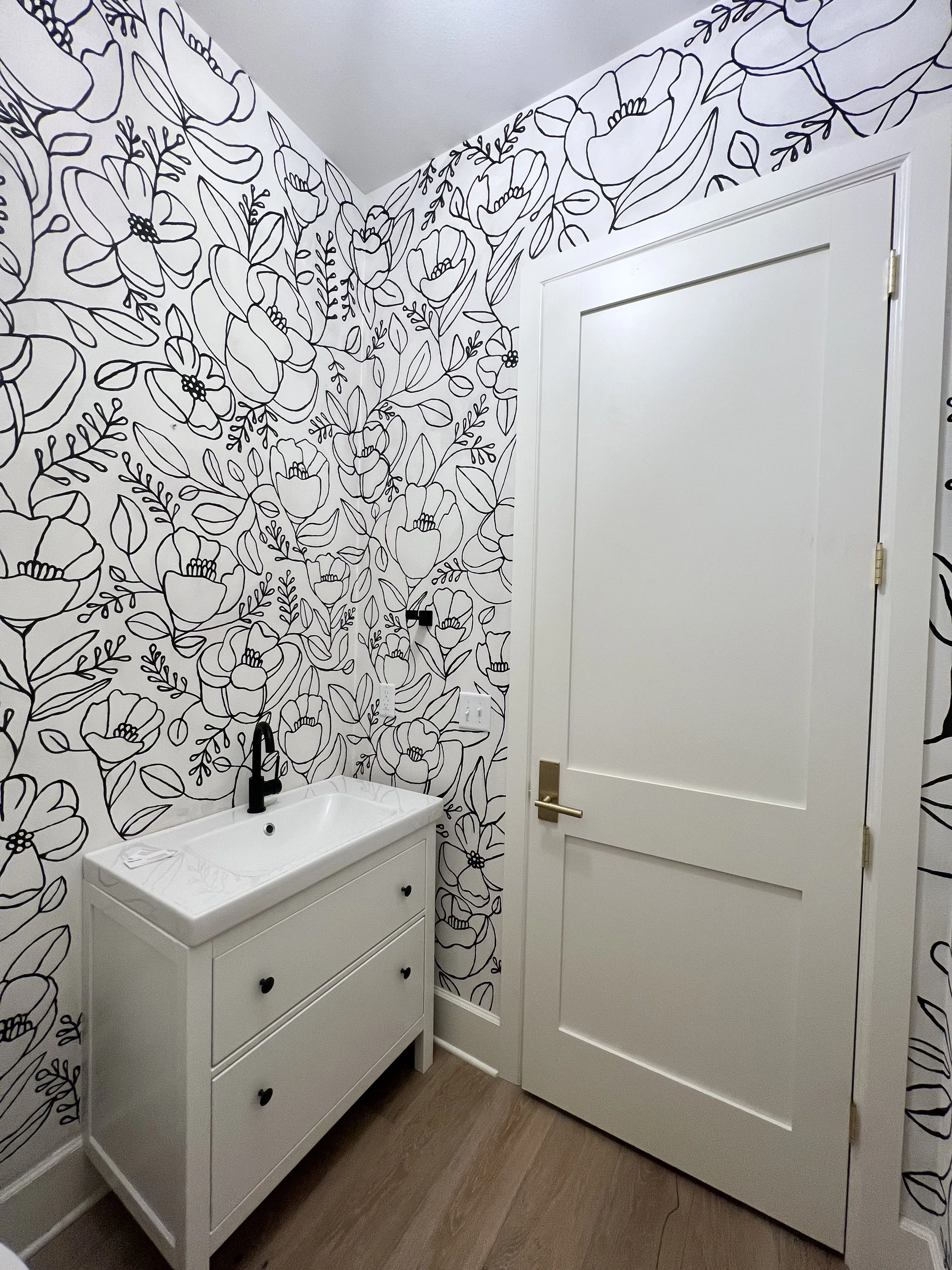 A small bathroom with floral wallpaper, a white vanity with a sink, a black faucet, and a closed white door.