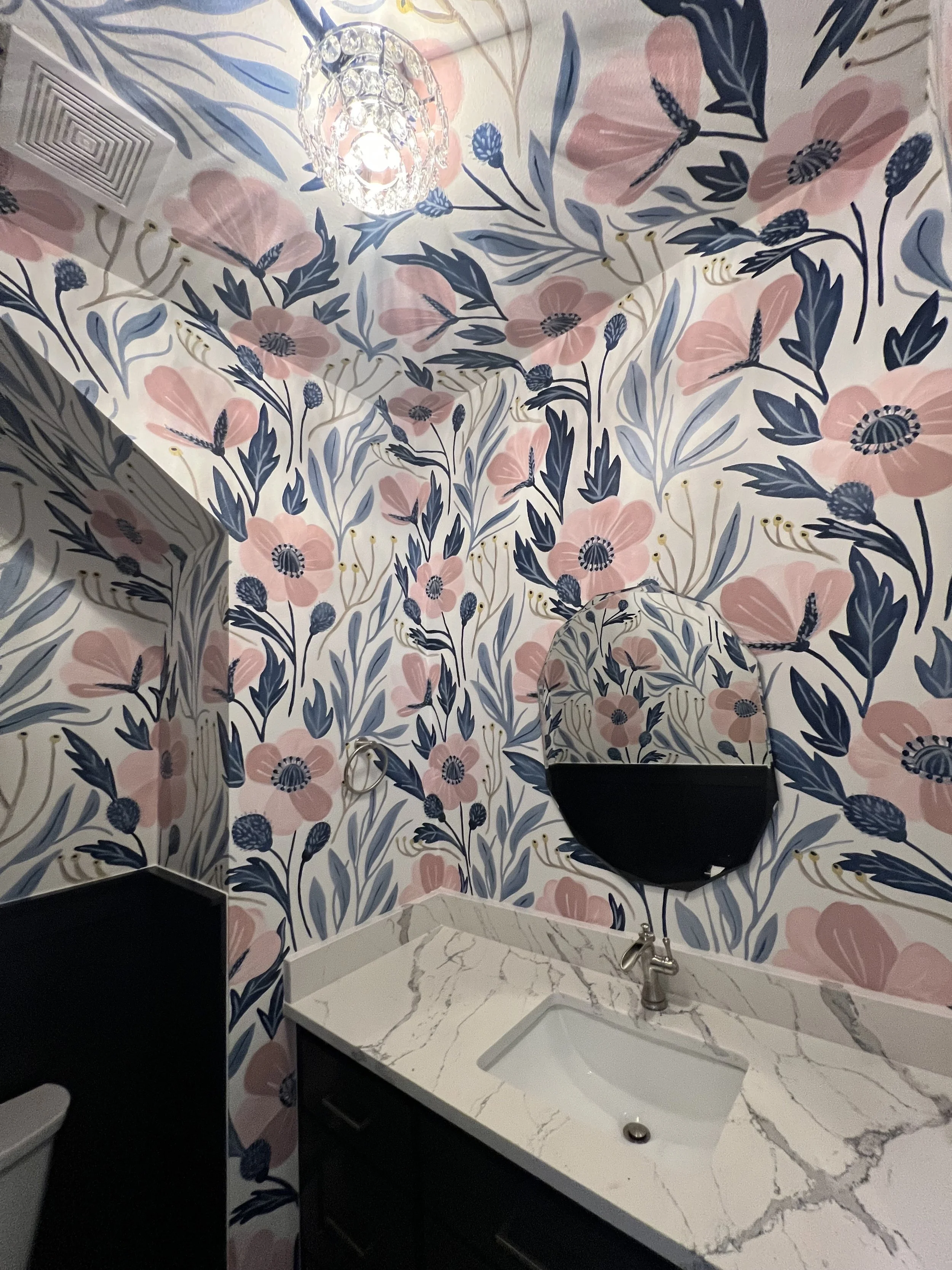 A bathroom with floral wallpaper in pink, blue, and white, a white marble countertop with a built-in sink, a round mirror, a chrome faucet, and a wall-mounted black shelf.