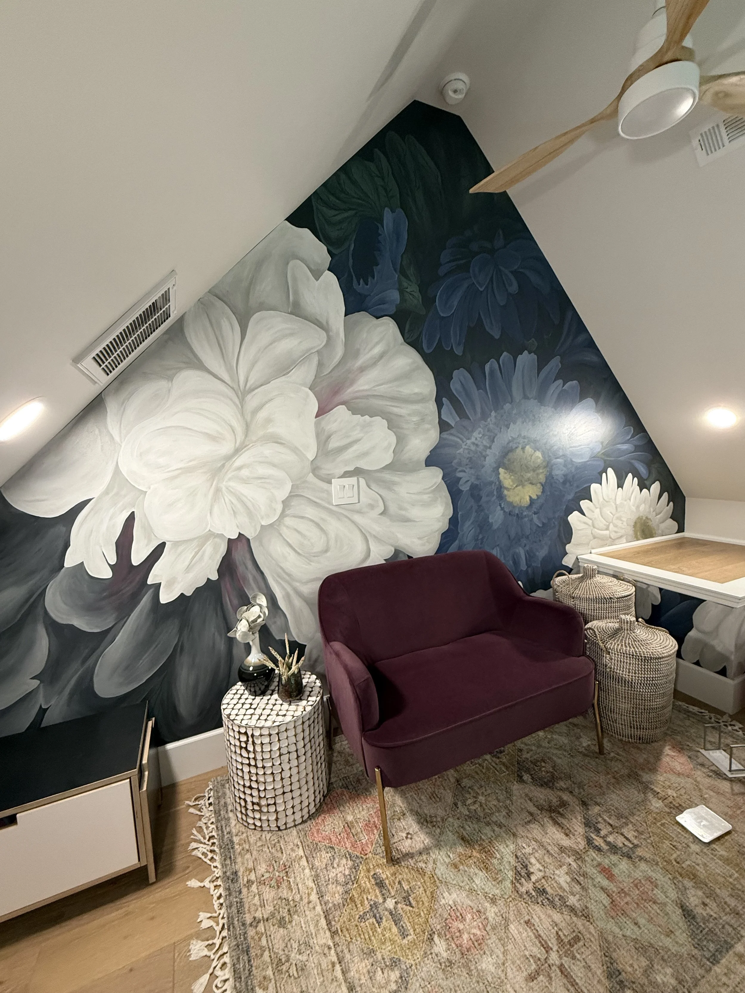 A room with a large floral mural featuring white, blue, and dark-colored flowers on an angular wall. The room contains a purple loveseat, a patterned side table with decorative items, and a woven basket. There is a radiator vent on the left side, and a ceiling fan with wooden blades overhead. The floor has a colorful, patterned rug.