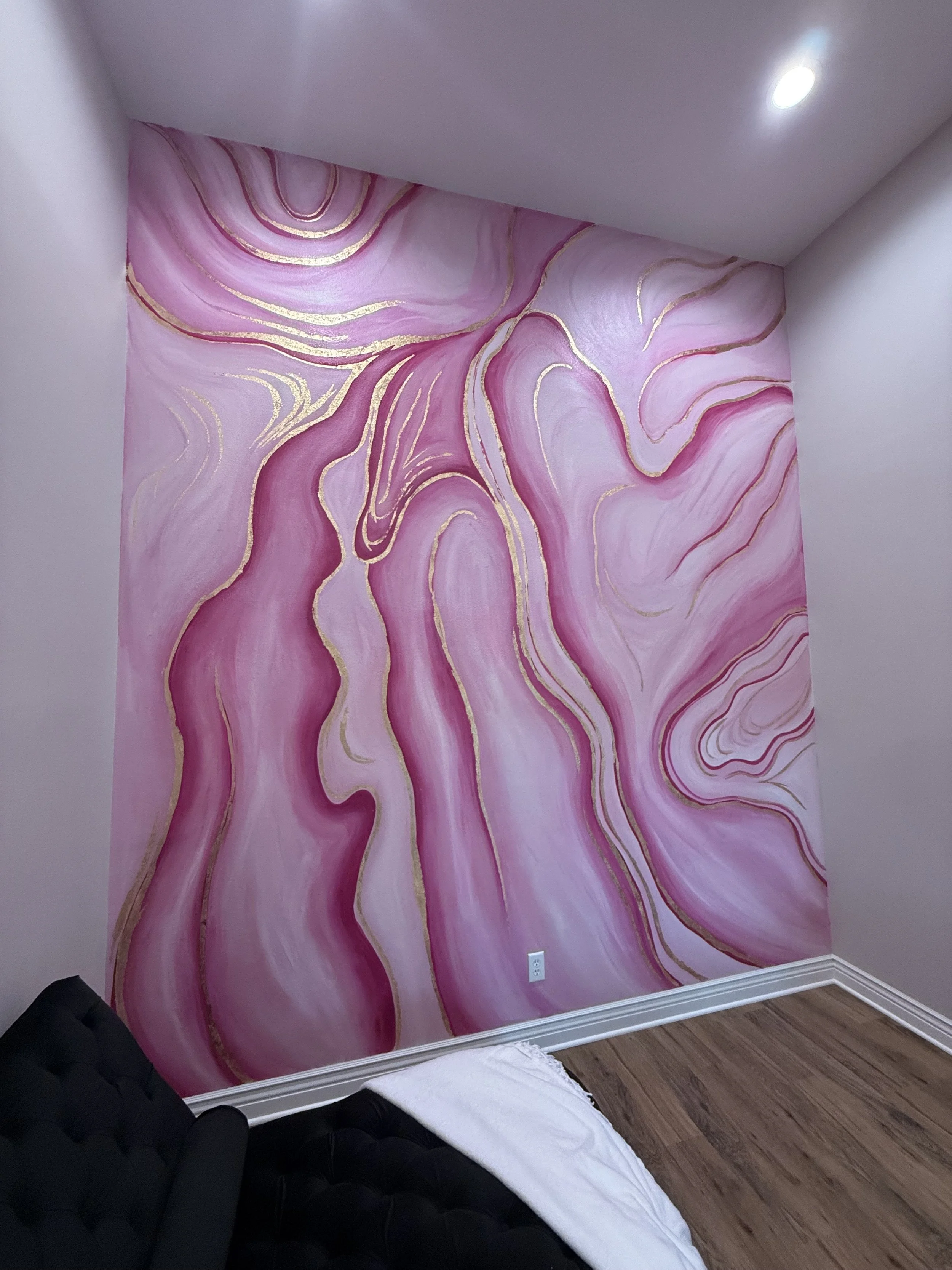 Pink abstract mural with gold accents on a corner wall in a room with a hardwood floor and a bed with black headboard and white bedding.
