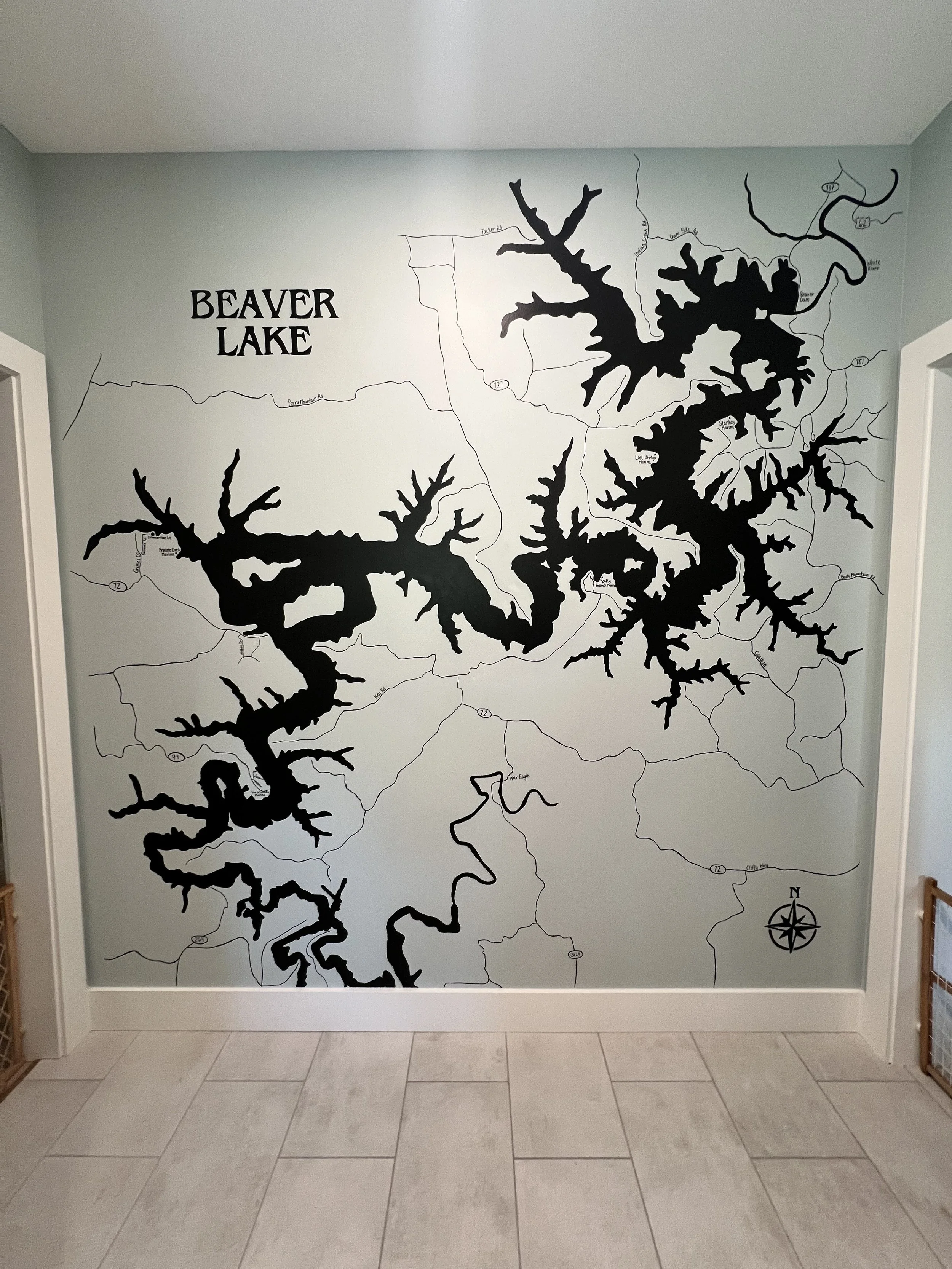 Black silhouette of Beaver Lake's shoreline and water body on a wall mural, with surrounding roads, a compass rose, and the text 'Beaver Lake' in the upper left corner.