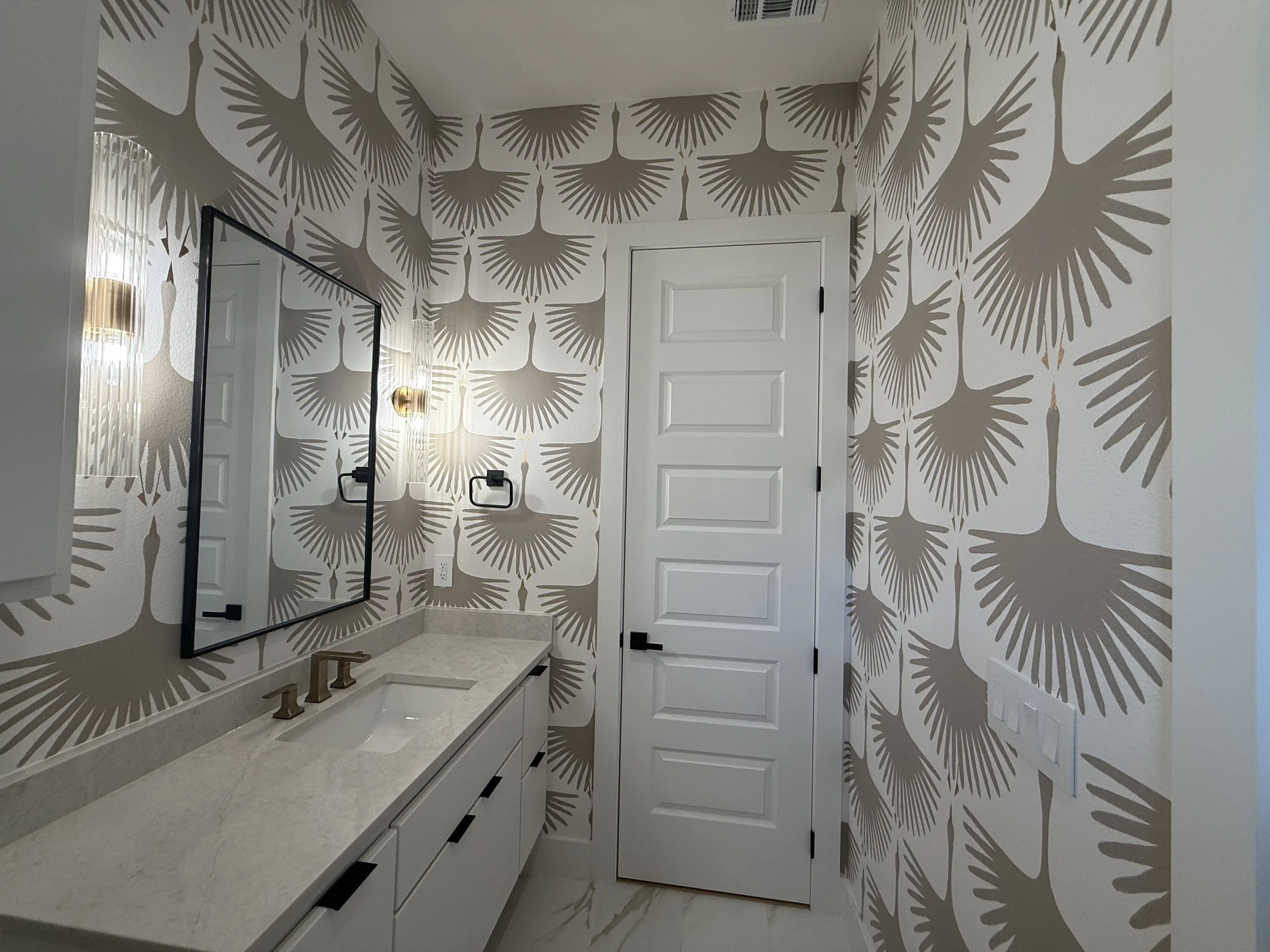 Bathroom with white vanity, marble countertop, single sink, gold fixtures, large mirror, wallpaper with large beige fan-like leaf pattern, white door, gold wall-mounted sconce, black towel hooks, multi-switch panel.