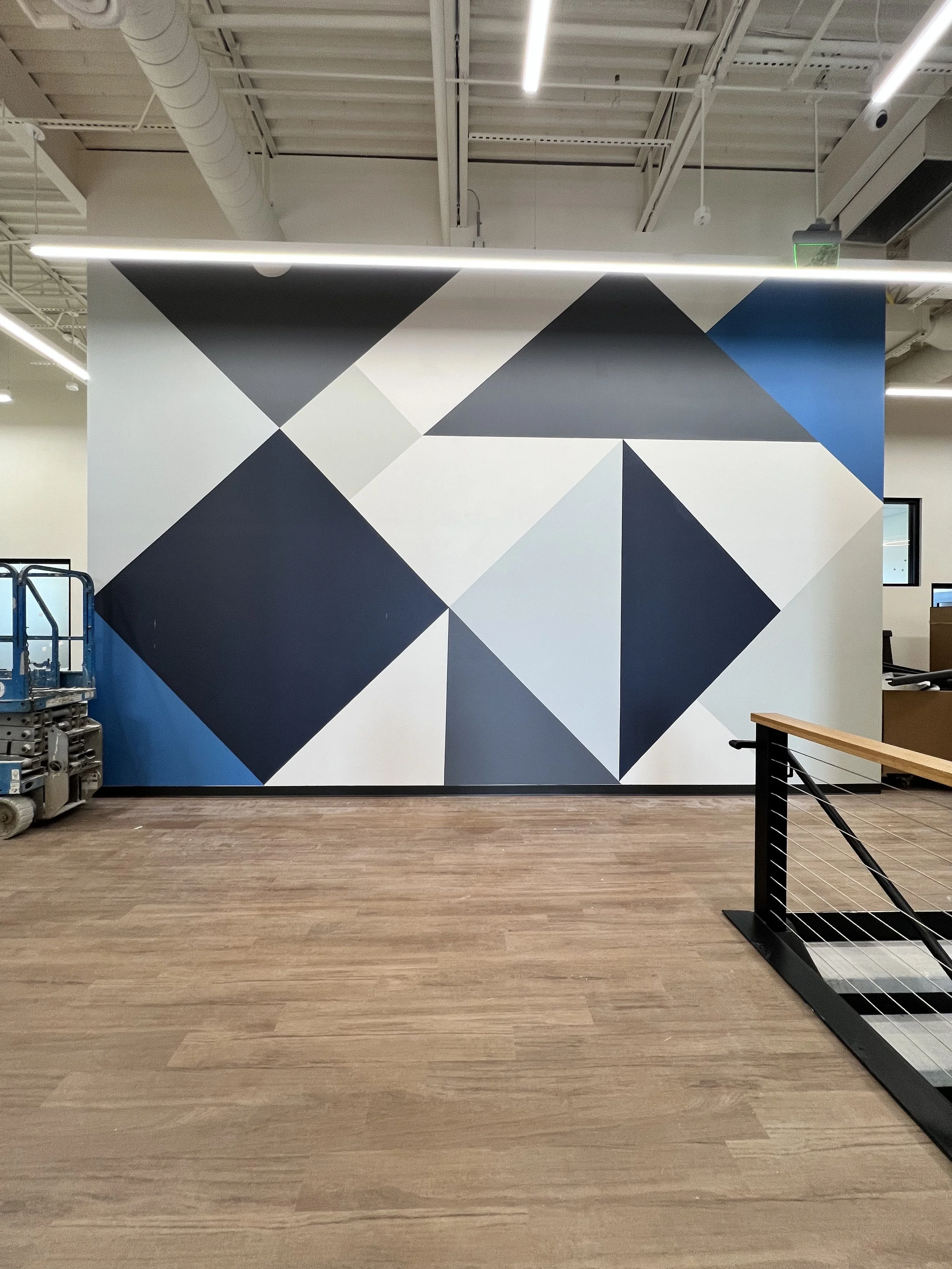Interior of a modern office with a large geometric mural wall in shades of blue, black, gray, and white, with a wooden floor and some office equipment on the left side.