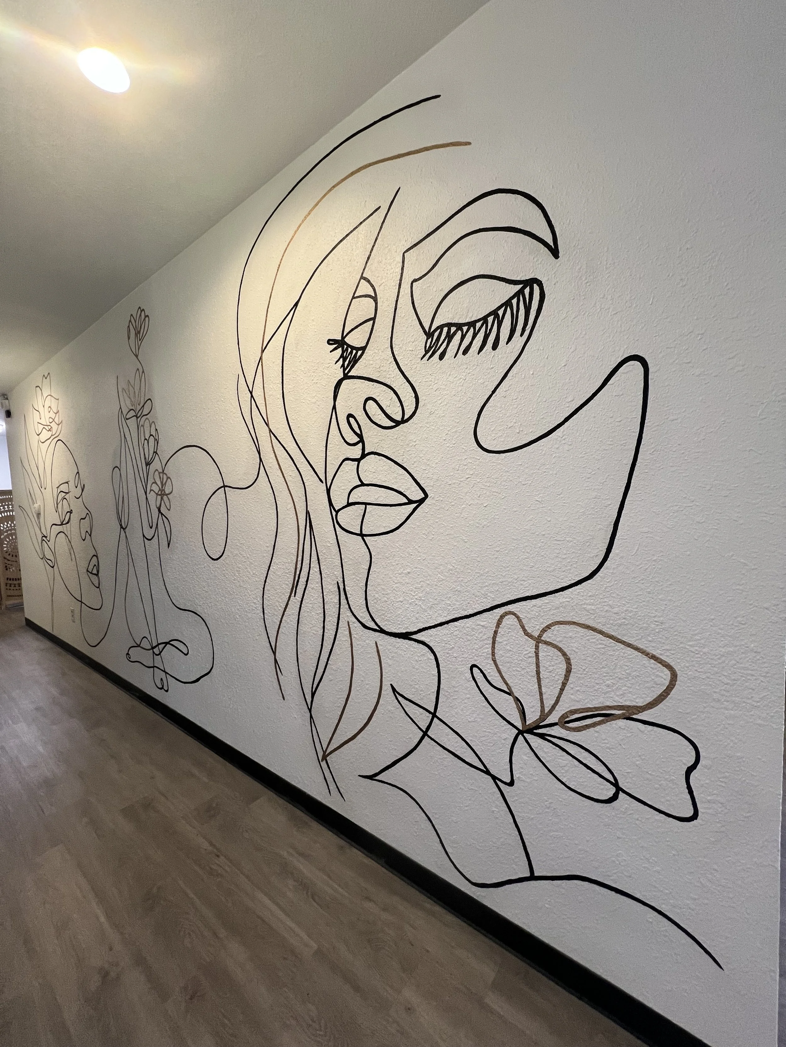 Wall art of a continuous line drawing of a woman's face with closed eyes, lips, and hair, on a white textured wall in an indoor space.