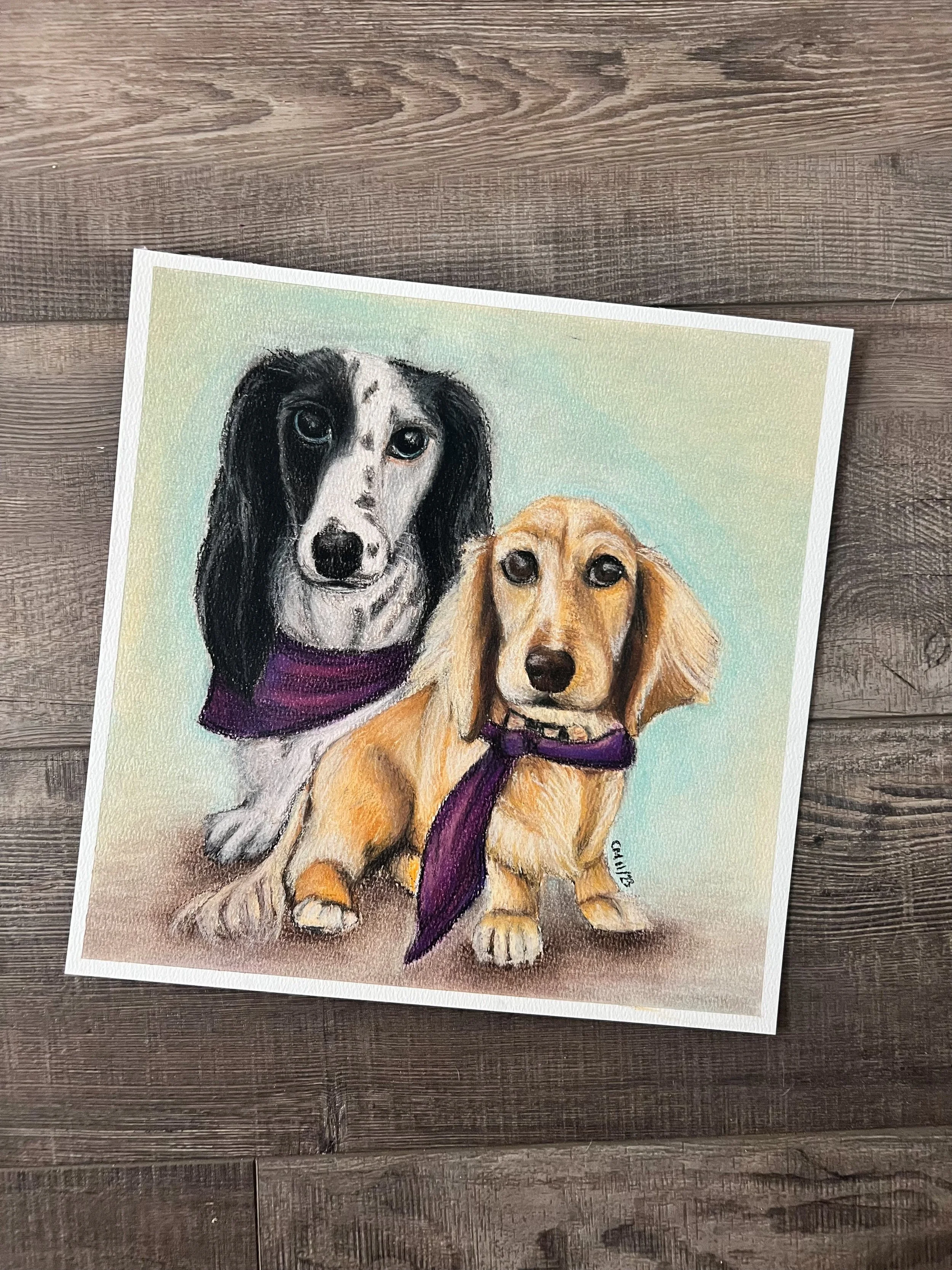 Colored drawing of two dogs, a black and white dog and a tan dog, both wearing purple bandanas, on a wooden surface background.