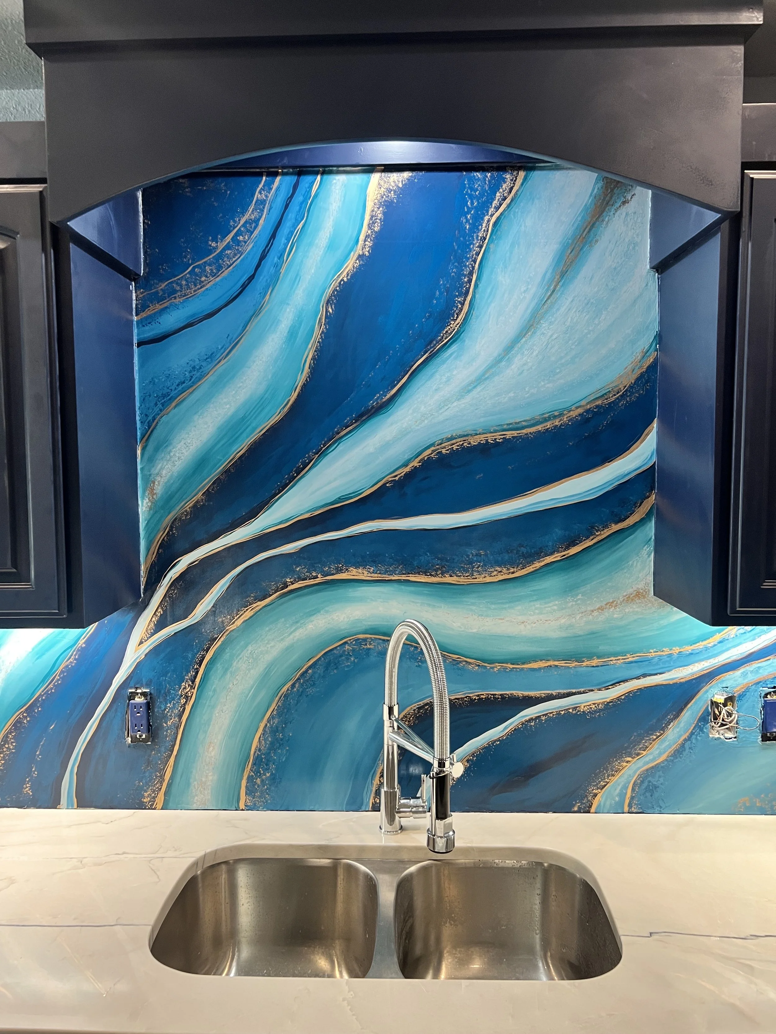 Kitchen backsplash with a blue and gold abstract swirl design, stainless steel sink and faucet, black cabinets.