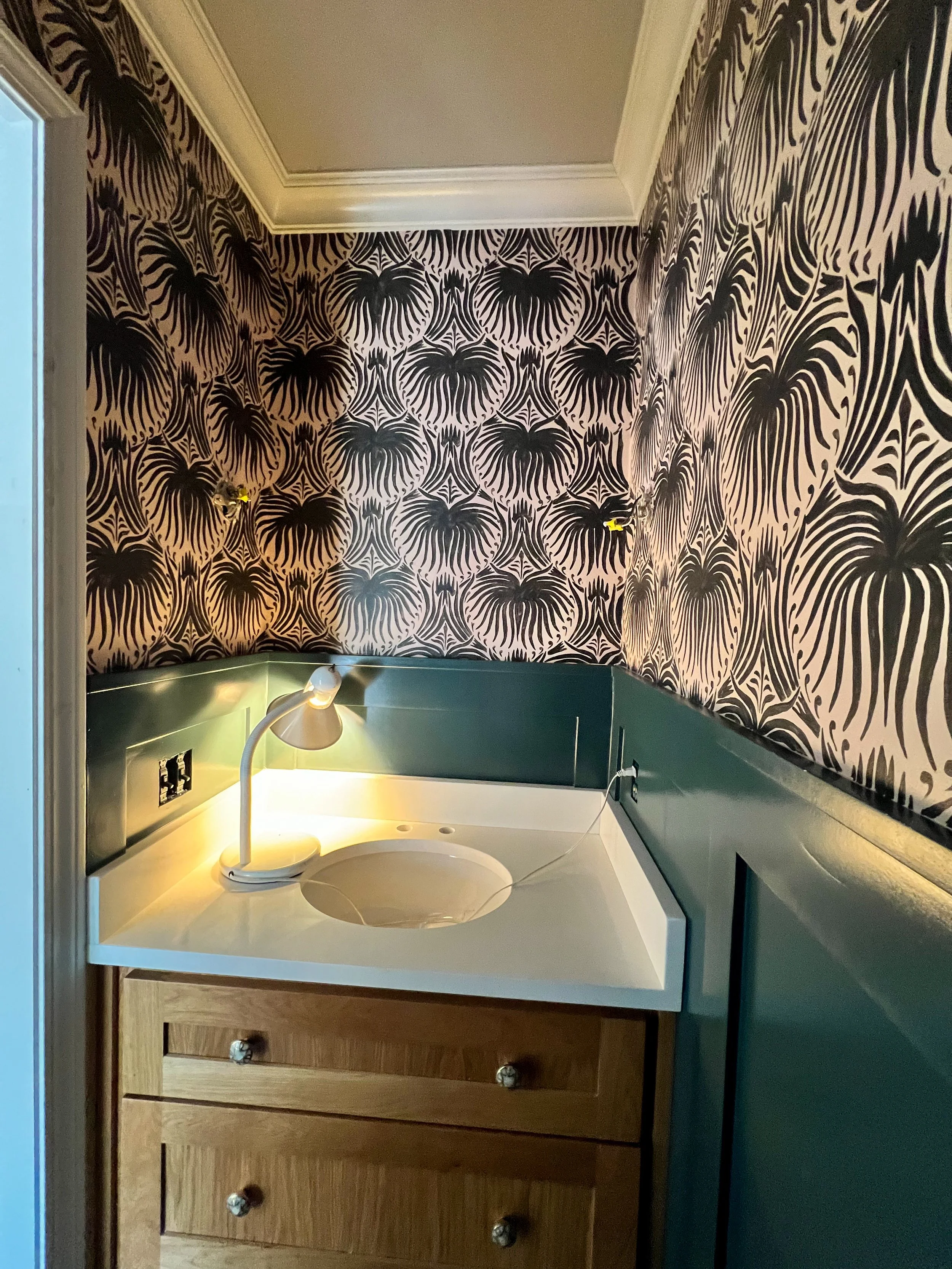 A bathroom vanity with a built-in sink and a flexible reading lamp on the left, under a green backsplash, against wallpaper with a black and beige tropical leaf pattern.