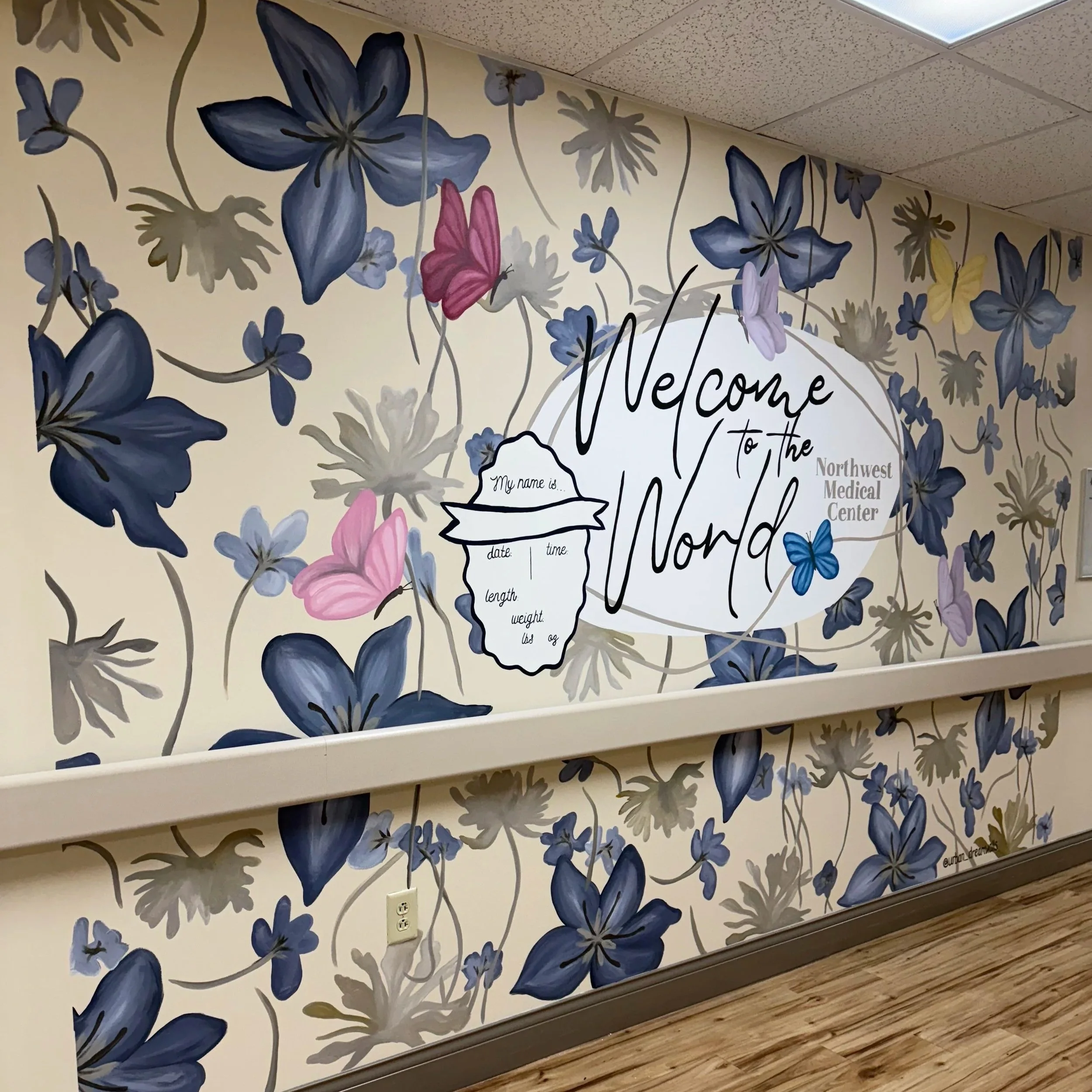 Decorative wall with blue and purple flowers and butterflies, featuring a sign that says 'Welcome to the Nord' at Northwest Medical Center with a form to fill in name, date, time, length, weight, and height.