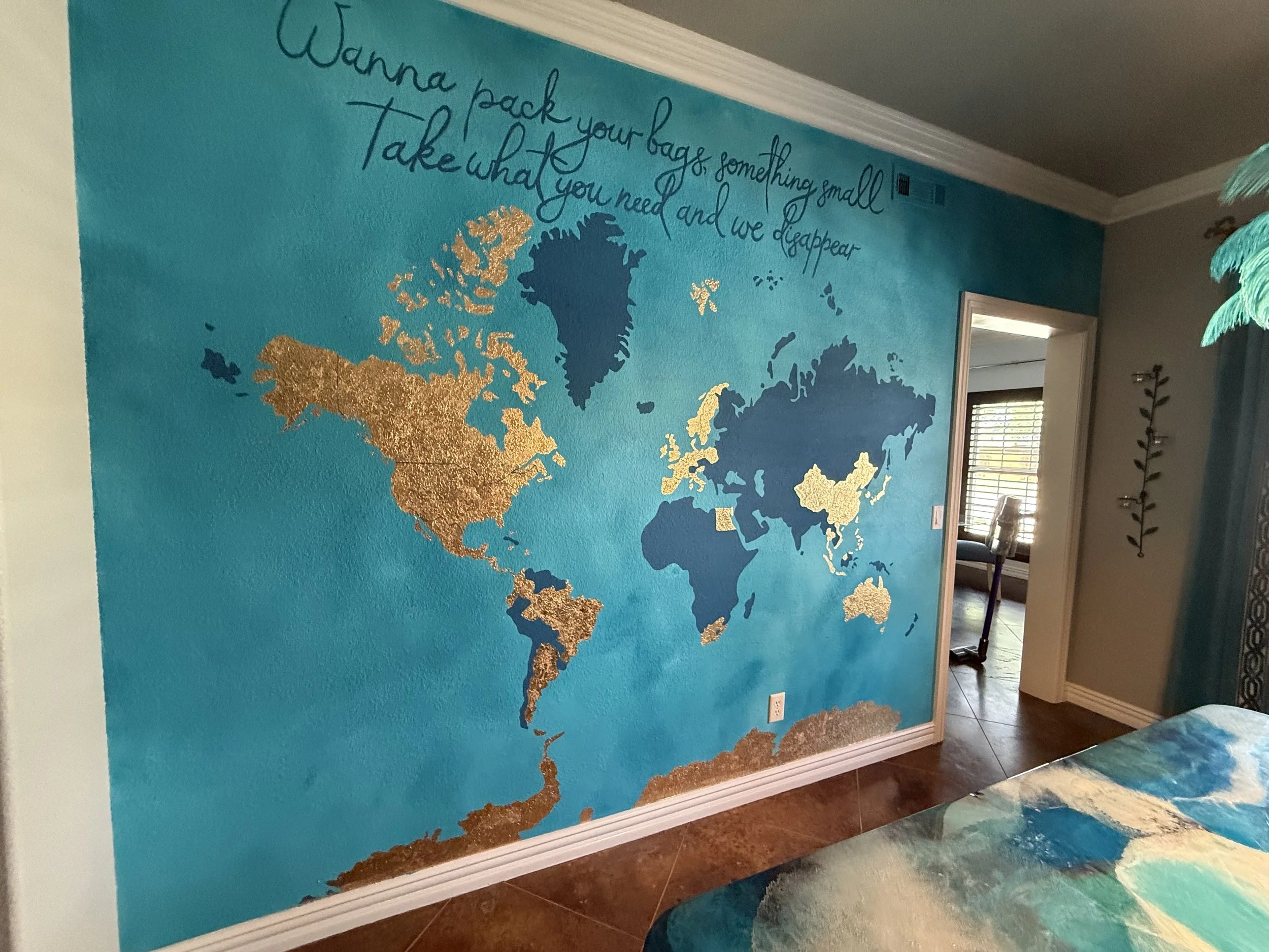 Decorative wall mural of a world map with gold and blue continents, featuring a quote written in cursive at the top that says, 'Wanna pack your bags, something small. Take what you need and we disappear.' The room has tiled flooring and a doorway leading to another room with window blinds.