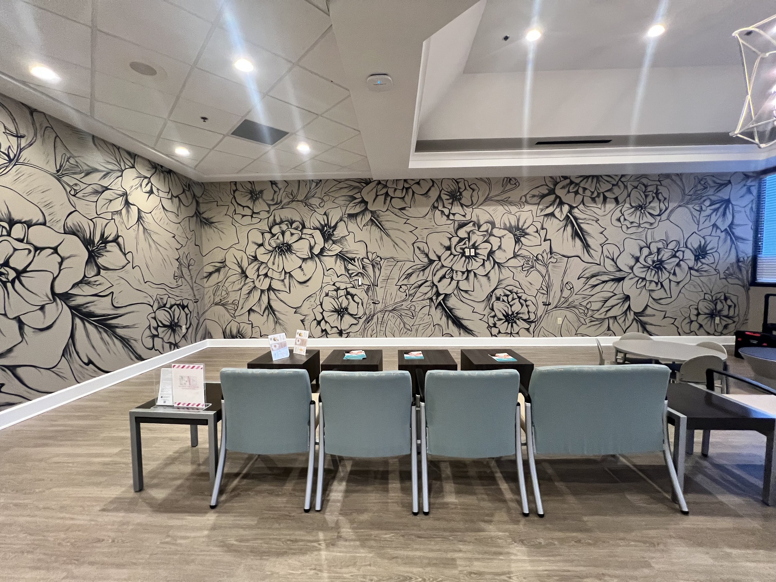An empty room with a floral mural on the wall, a row of six blue chairs in front, and three small black tables with pamphlets on them. There is a window with blue blinds on the right side.
