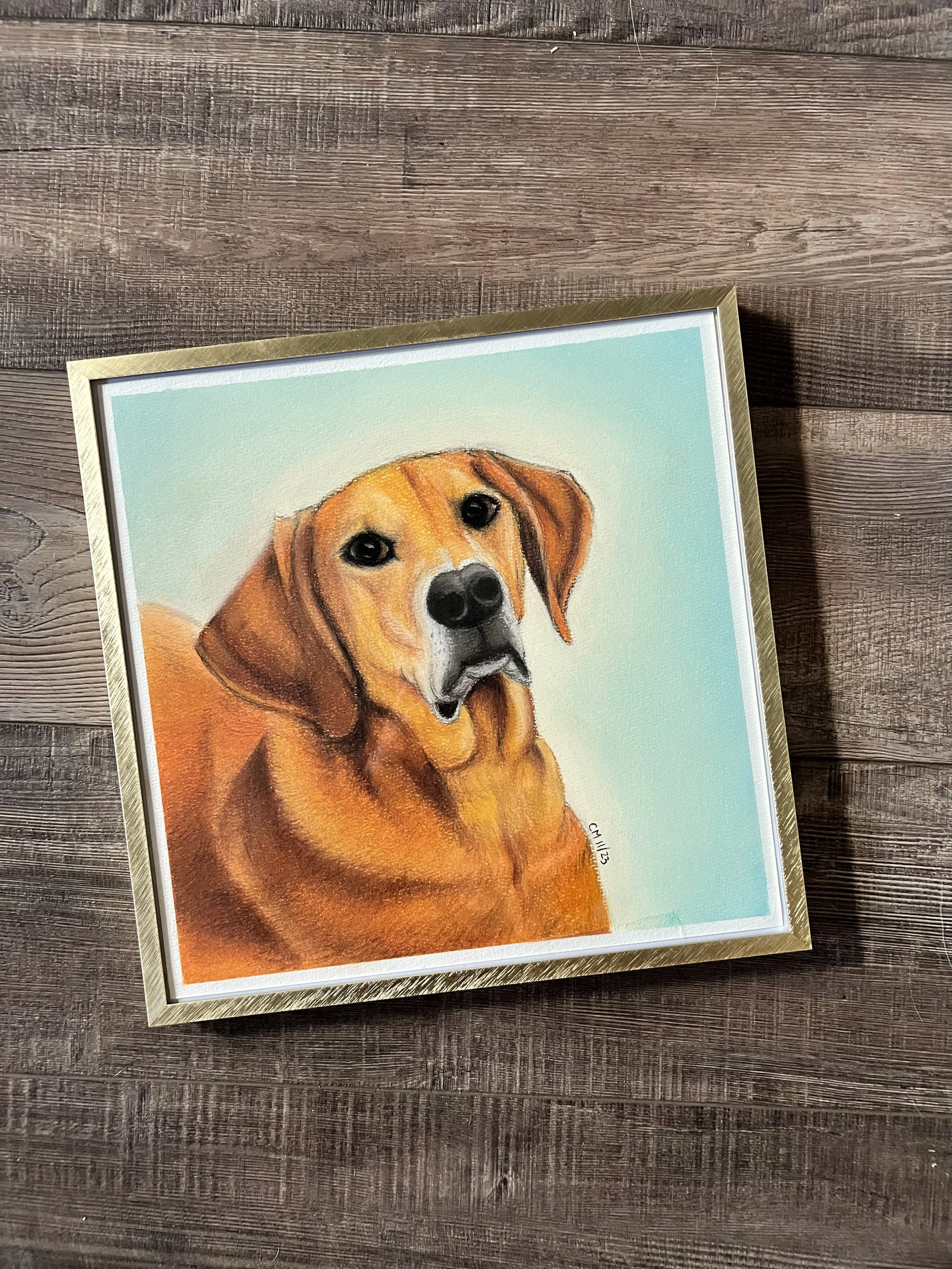 Framed painting of a dog, likely a retriever, with a light blue background, placed on a wooden surface.
