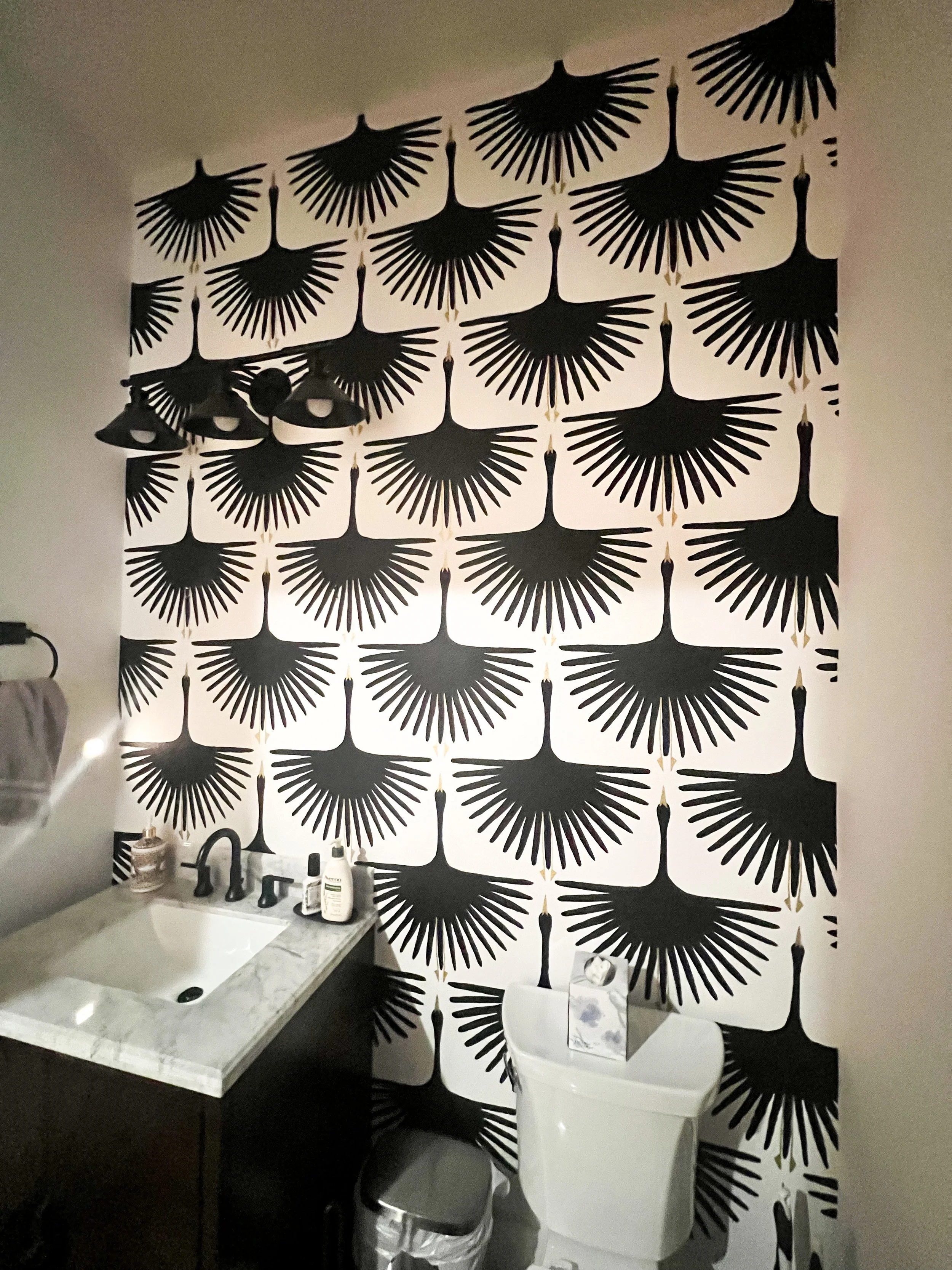 Bathroom with black and white patterned wallpaper featuring geometric fan shapes, a small sink with soap and skincare products, a toilet with a tissue box on top, a trash bin, and three ceiling lights.