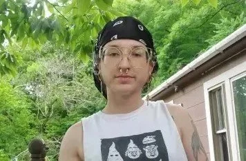 A white nonbinary person with a silver septum ring in a white muscle tee and black bandana.
