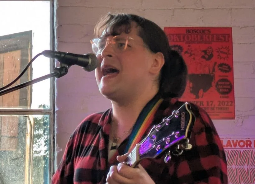 A white trans woman wearing a red buffalo plaid shirt and playing electric guitar and singing. 