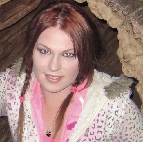 A white trans woman with red hair in two braids and a cheetah print jacket smiles into the camera.