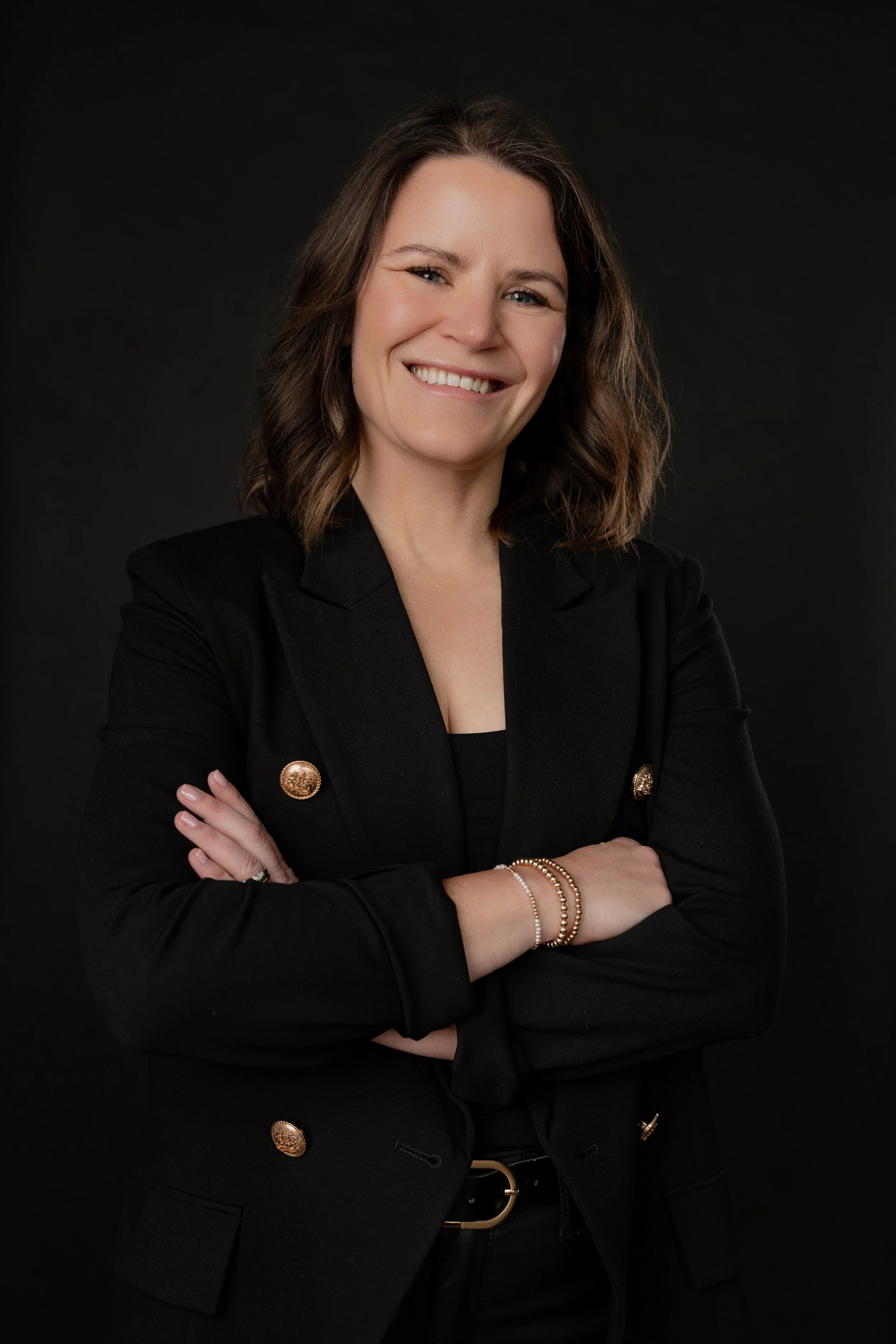 Millie Sessions, director of client experience at Insula Aesthetics, smiling with arms crossed, wearing a black blazer against a dark studio background.