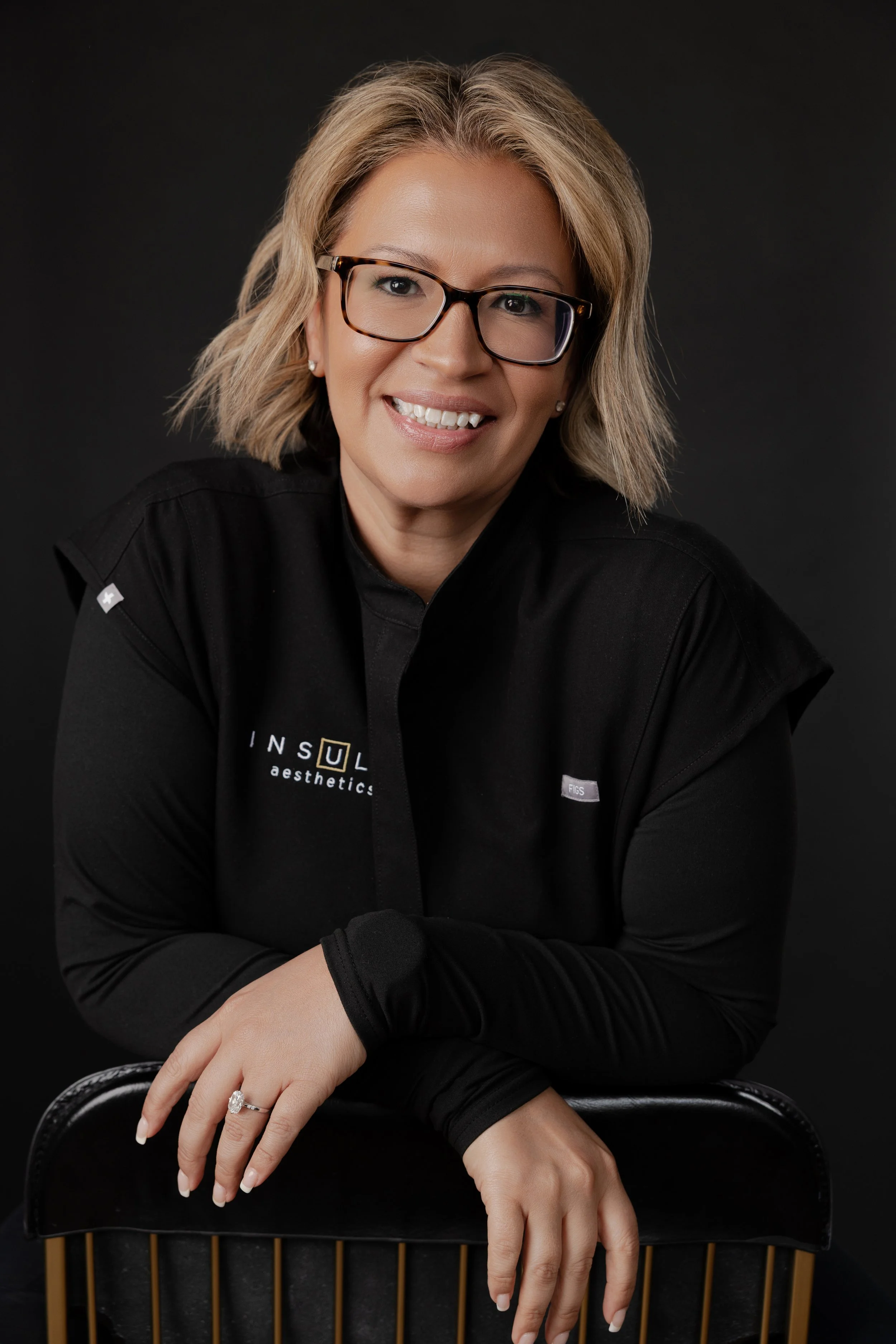 Jen Reitz, front desk coordinator at Insula Aesthetics, smiling and wearing glasses in a black branded top, resting arms on a chair against a dark studio background.
