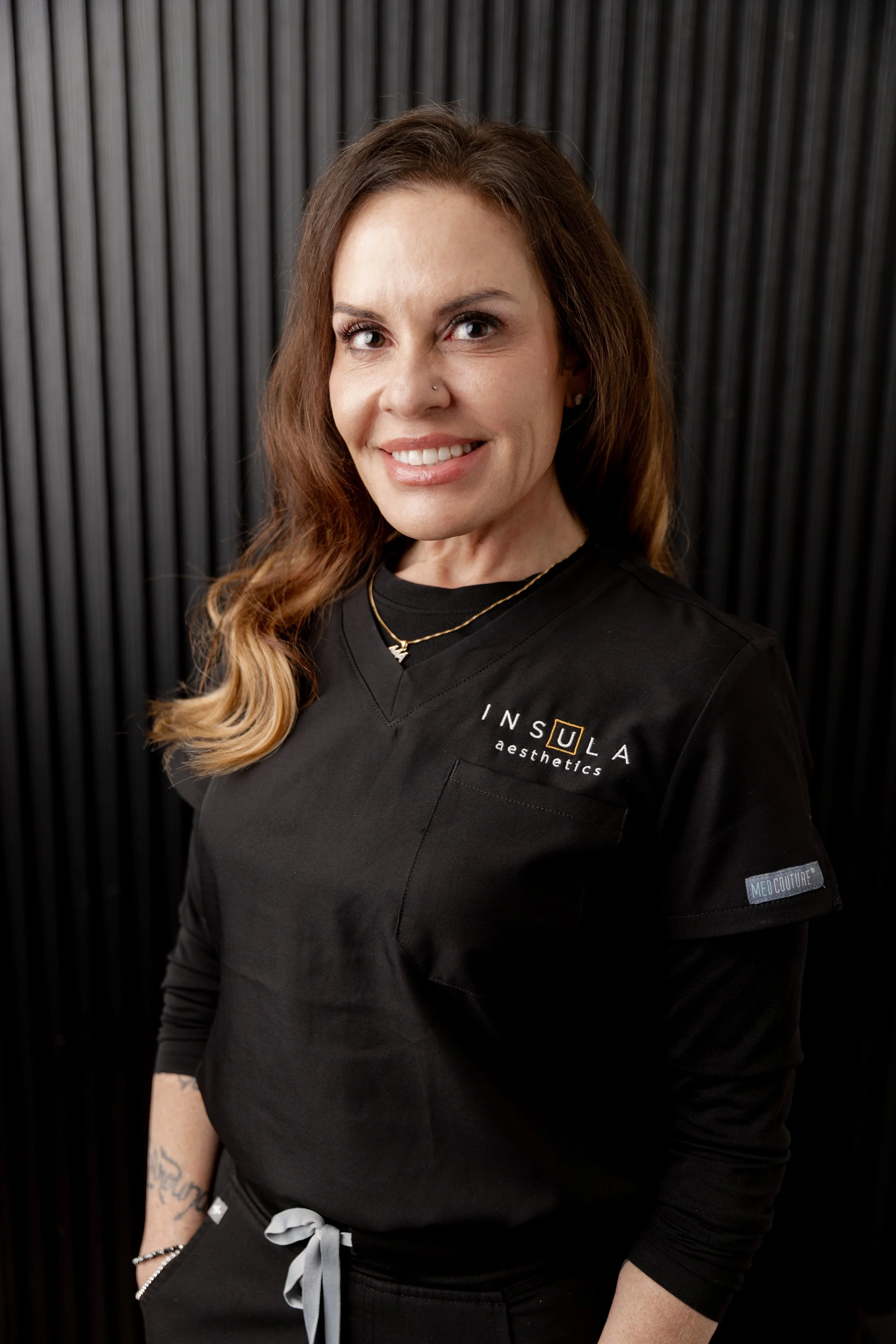 Joanna Bentz, LE, licensed esthetician at Insula Aesthetics, smiling in a black branded scrub top against a ribbed dark studio backdrop.