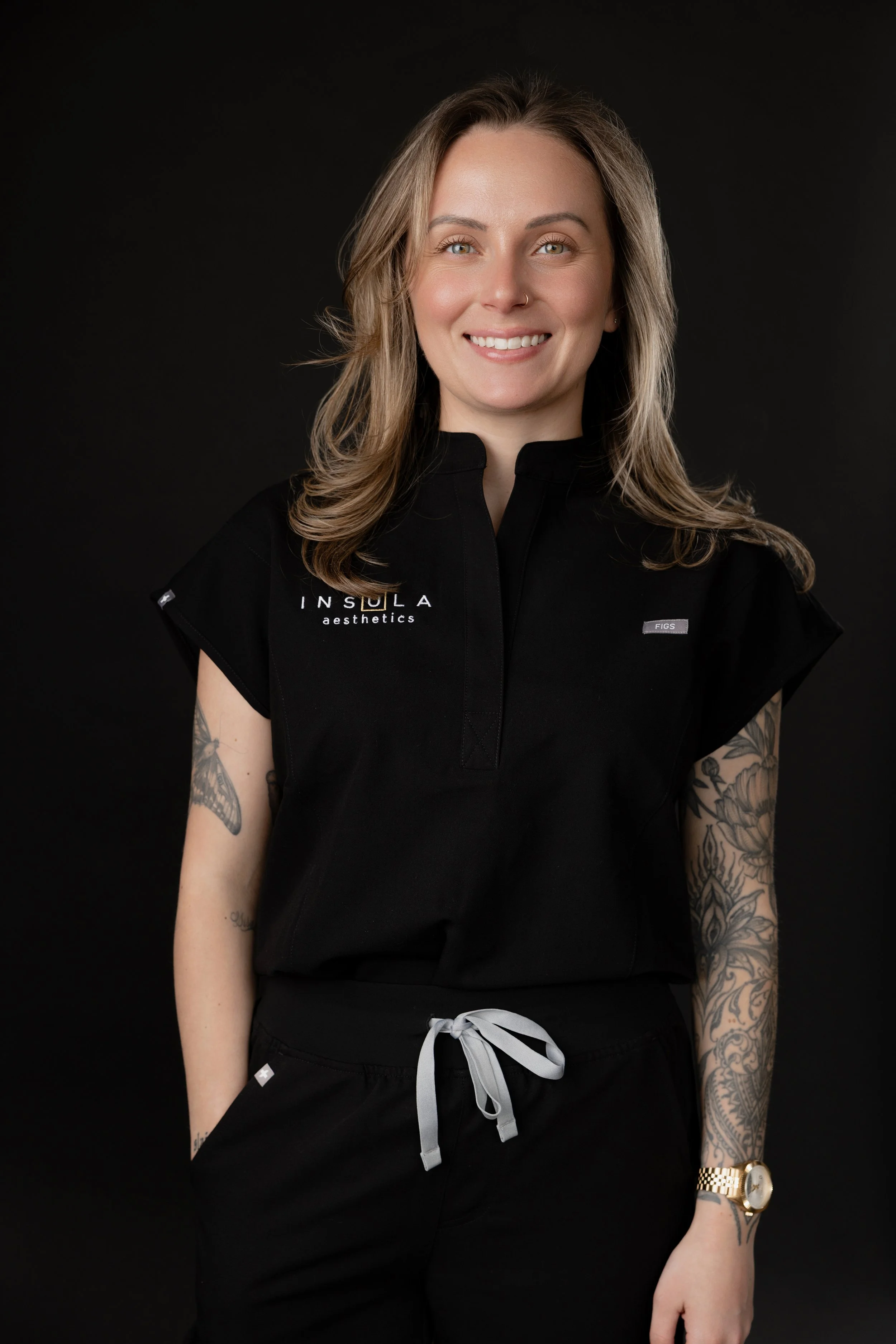 Brenna Bell, director of operations at Insula Aesthetics, smiling in a black branded polo, standing against a dark studio background.